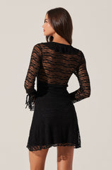 A woman is wearing a black, sheer, long-sleeved dress with a textured, striped pattern. The dress features ruffled details on the shoulders and tied fabric accents on the sleeves. The skirt portion of the dress is slightly flared and has a matching textured pattern. The fabric appears lightweight and see-through, revealing the skin underneath.