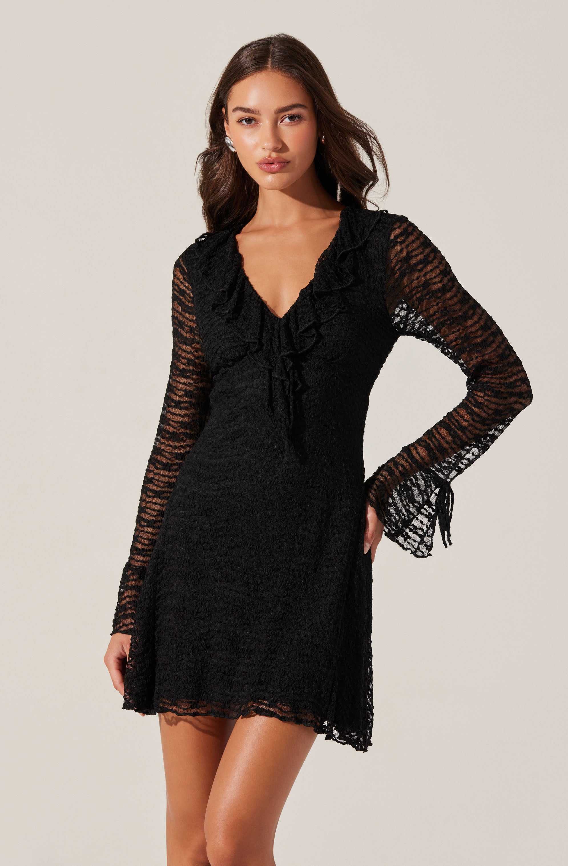 A woman is wearing a black dress made of textured, semi-sheer fabric. The dress features long, transparent lace sleeves with a ruched detail at the cuffs, a V-neckline, and a ruffled collar that extends down the front.