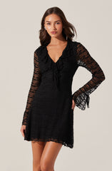A woman is wearing a black dress made of textured, semi-sheer fabric. The dress features long, transparent lace sleeves with a ruched detail at the cuffs, a V-neckline, and a ruffled collar that extends down the front.