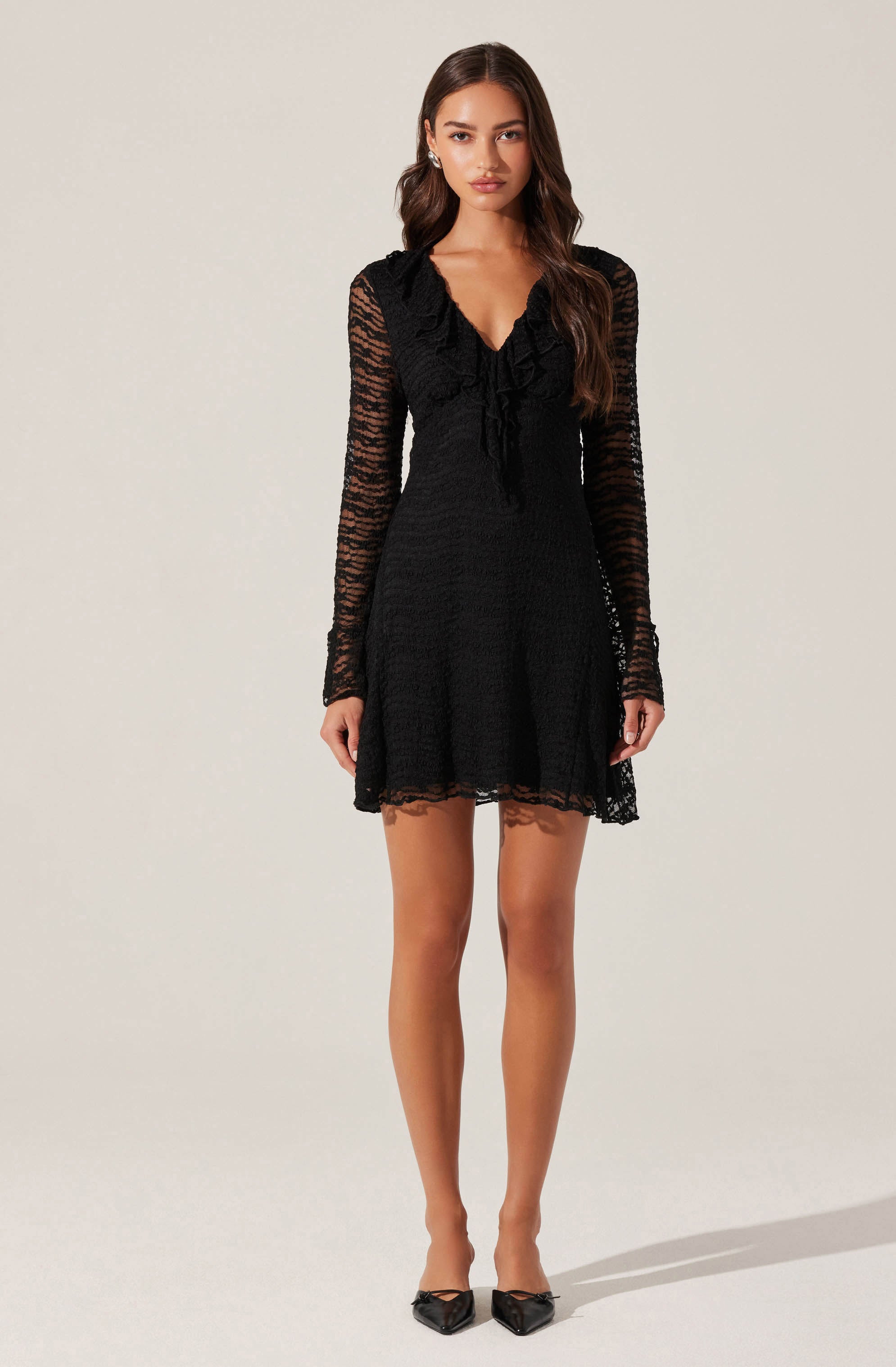 A woman is wearing a black long-sleeve dress made of sheer, lace-like fabric with a textured pattern. The dress features ruffle details along the shoulders and a V-neckline with a bow tie at the front. The hem of the dress is above the knees. She is also wearing black pointed-toe shoes with bows on the front.
