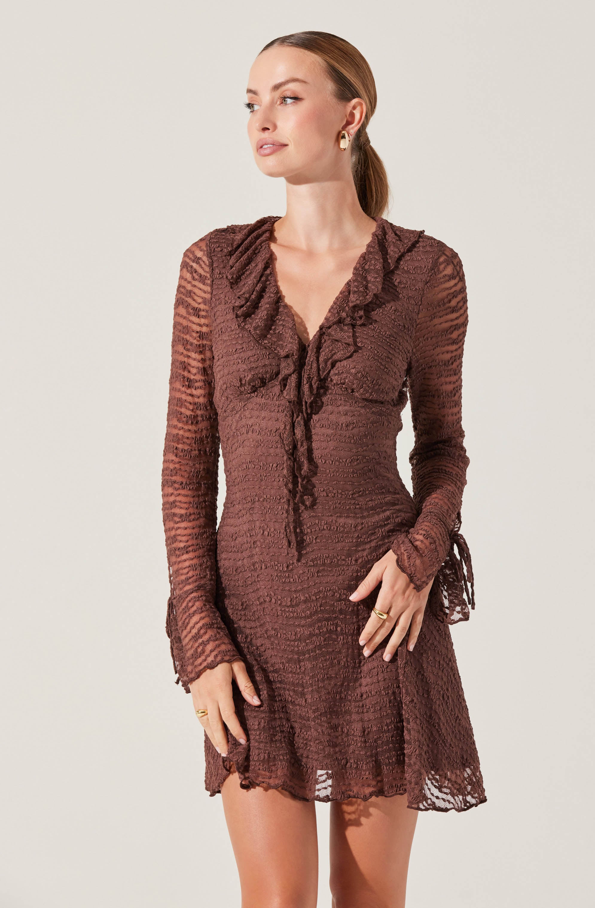 A woman is wearing a long-sleeved, semi-sheer dress made of textured, brown lace fabric. The dress features a ruffled collar that extends down the front and is tied at the waist with a matching lace ribbon. The sleeves have ruffled cuffs with lace ties, and the skirt is slightly flared with a scalloped hem.