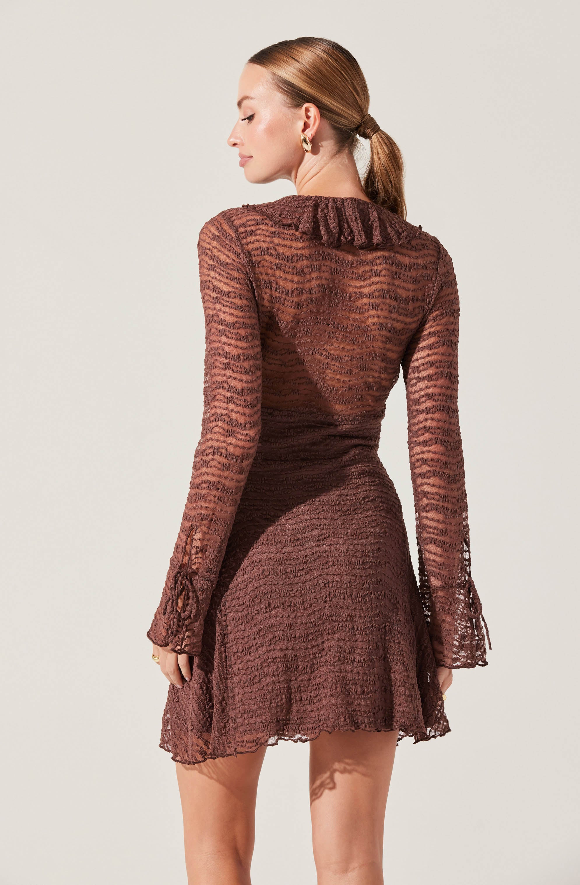 A woman is wearing a sheer, long-sleeved brown dress with a textured, zebra-like pattern. The dress features a ruffled collar and flared, ruffled cuffs on the sleeves. The fabric is see-through, revealing the outline of her body underneath.