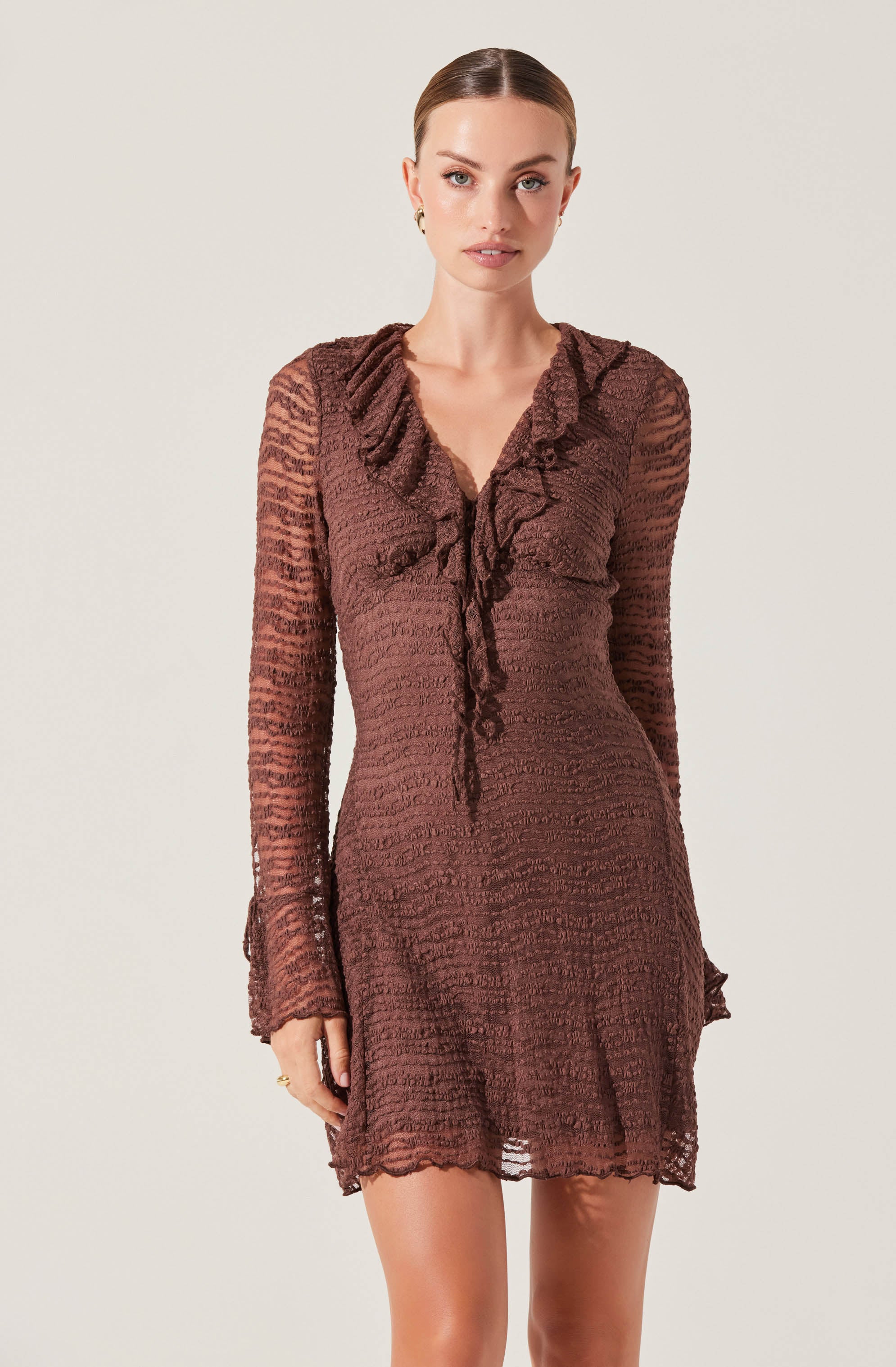 A woman is wearing a brown, textured, long-sleeved dress with a ruffled neckline that has a tie detail in the front. The dress has a semi-sheer appearance with a slightly ruffled hem at the bottom.