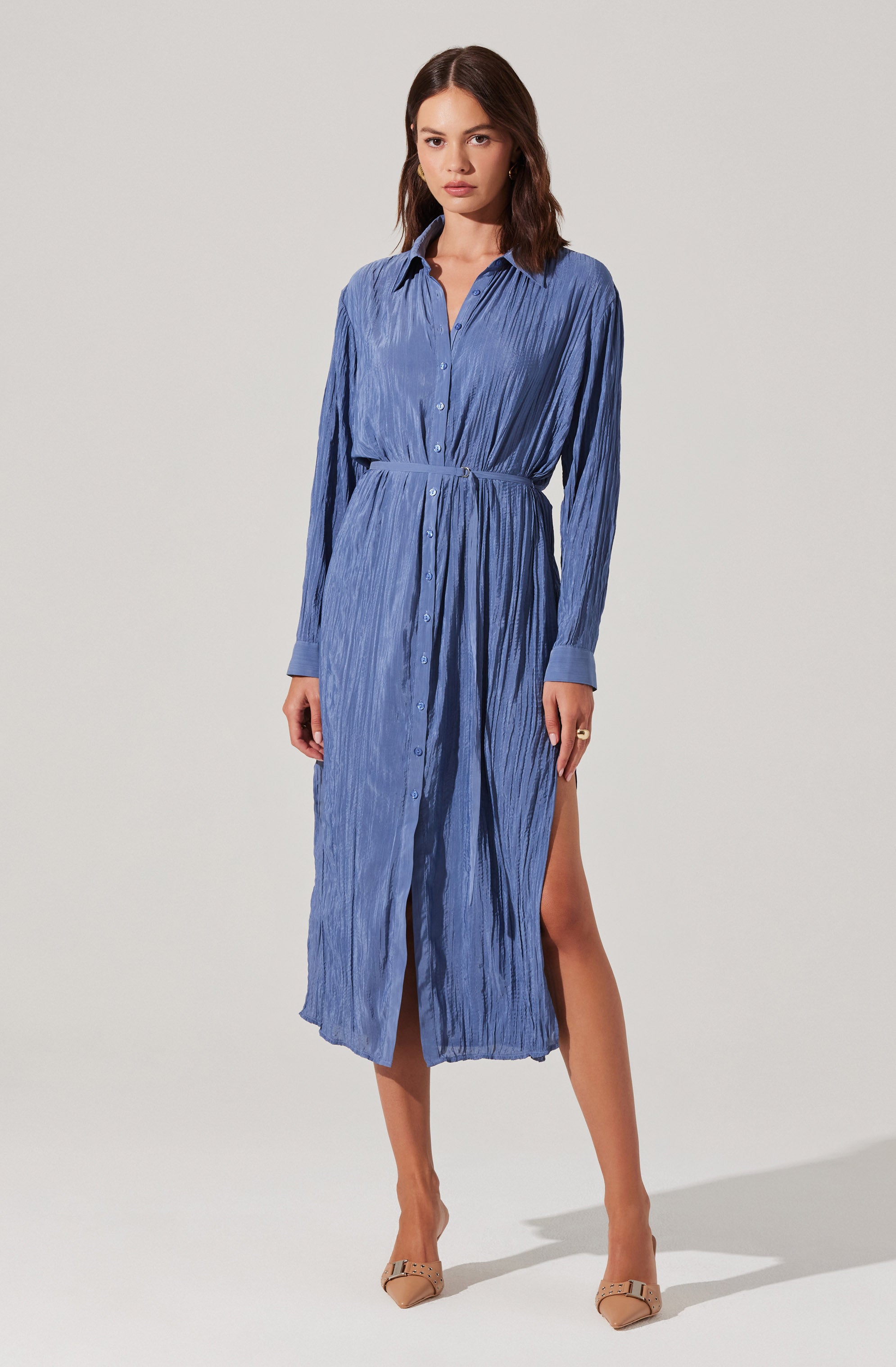 A woman is wearing a long, button-down dress made of lightweight, crinkled fabric in a medium blue color. The dress features a collar, long sleeves with cuffs, and a high slit on one side, revealing part of her leg.