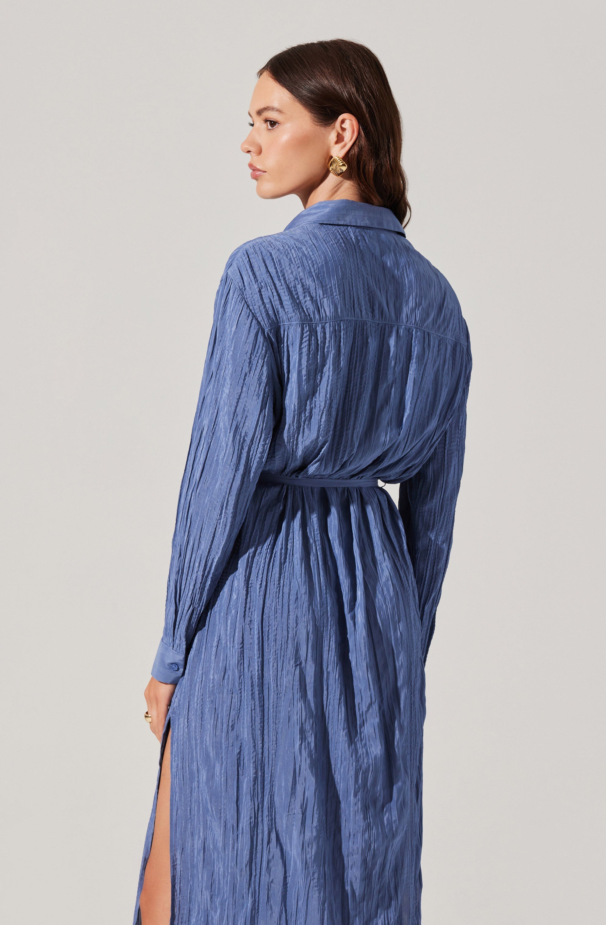 A woman is wearing a long, blue, textured dress with a collar and long sleeves. The dress has a gathered design at the waist with a matching fabric belt and features a high slit on one side, revealing part of her leg. She is accessorized with large gold earrings.