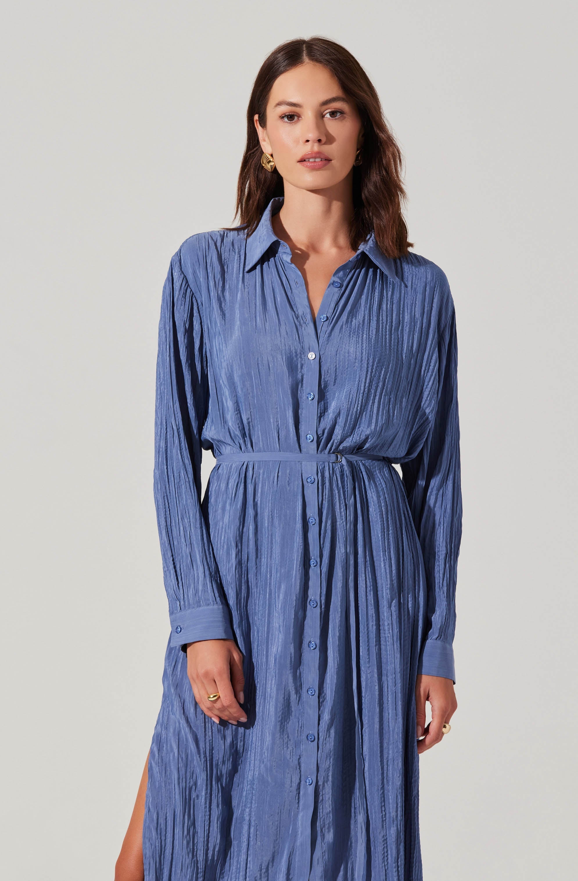 A woman is wearing a long, button-up shirt dress in a textured, light blue fabric with vertical pleats. The dress features a collar, long sleeves with cuff buttons, and a waist tie that cinches the dress at the waist. The dress has a slit on one side near the hem.
