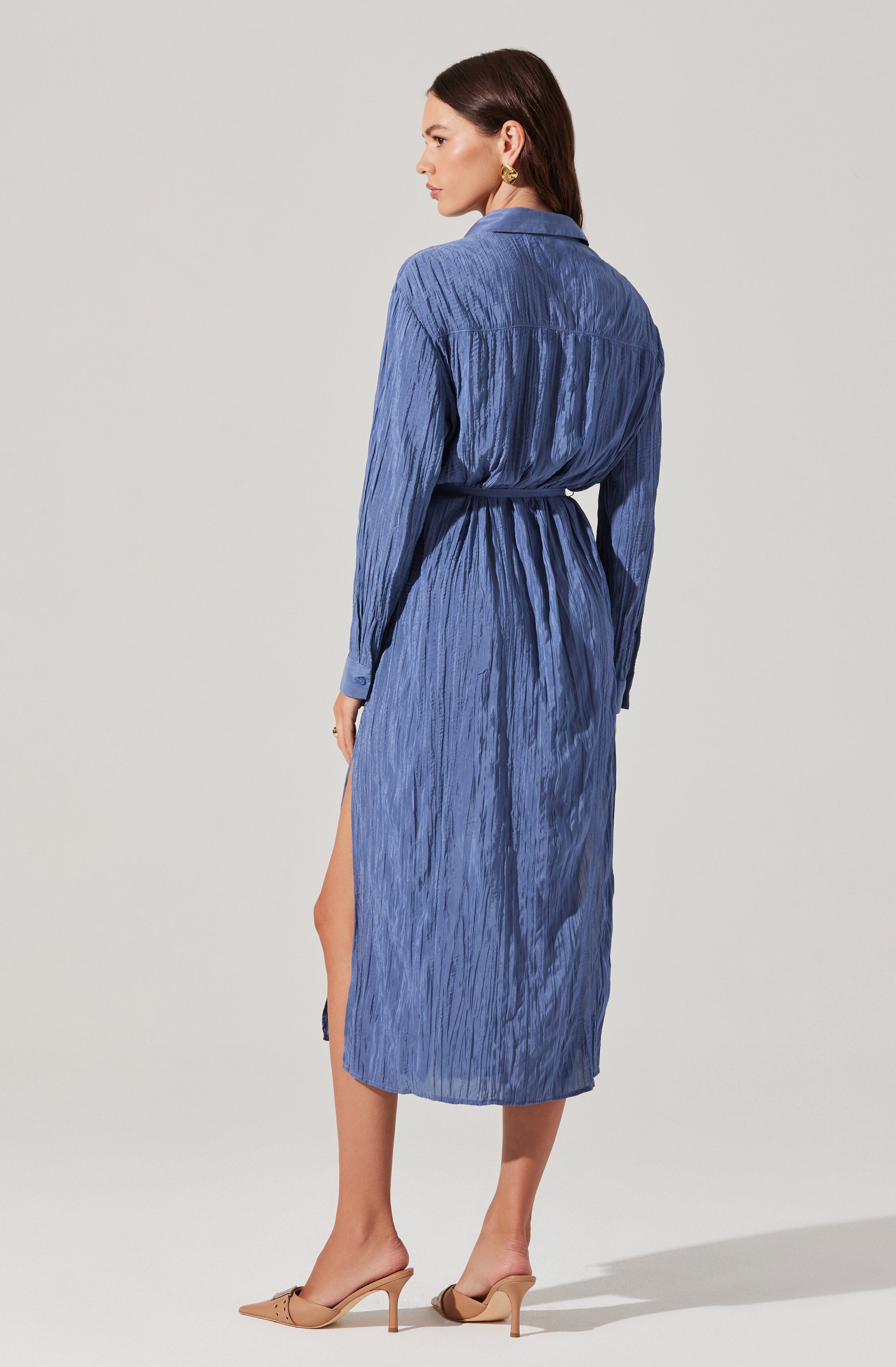 A woman is wearing a long, textured, blue dress with a collared neckline and long sleeves. The dress features a slit on the side, revealing part of her leg. She has a belt made from the same fabric tied around her waist. She completes the look with beige high-heeled shoes and gold earrings.