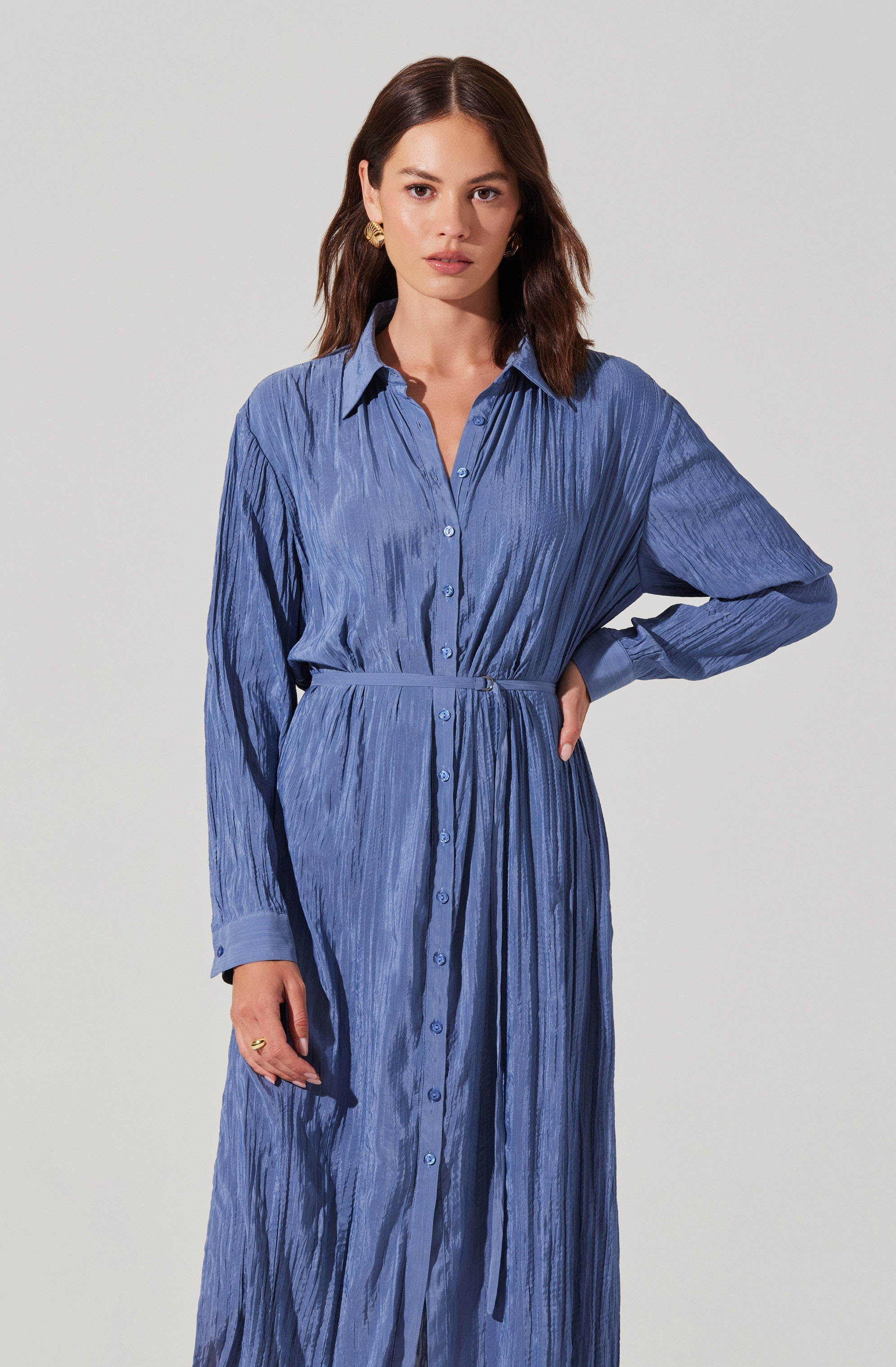A woman is wearing a long, button-up dress made of a lightweight, textured fabric in a shade of blue. The dress features a collar, long sleeves with cuffs, and a matching belt at the waist.