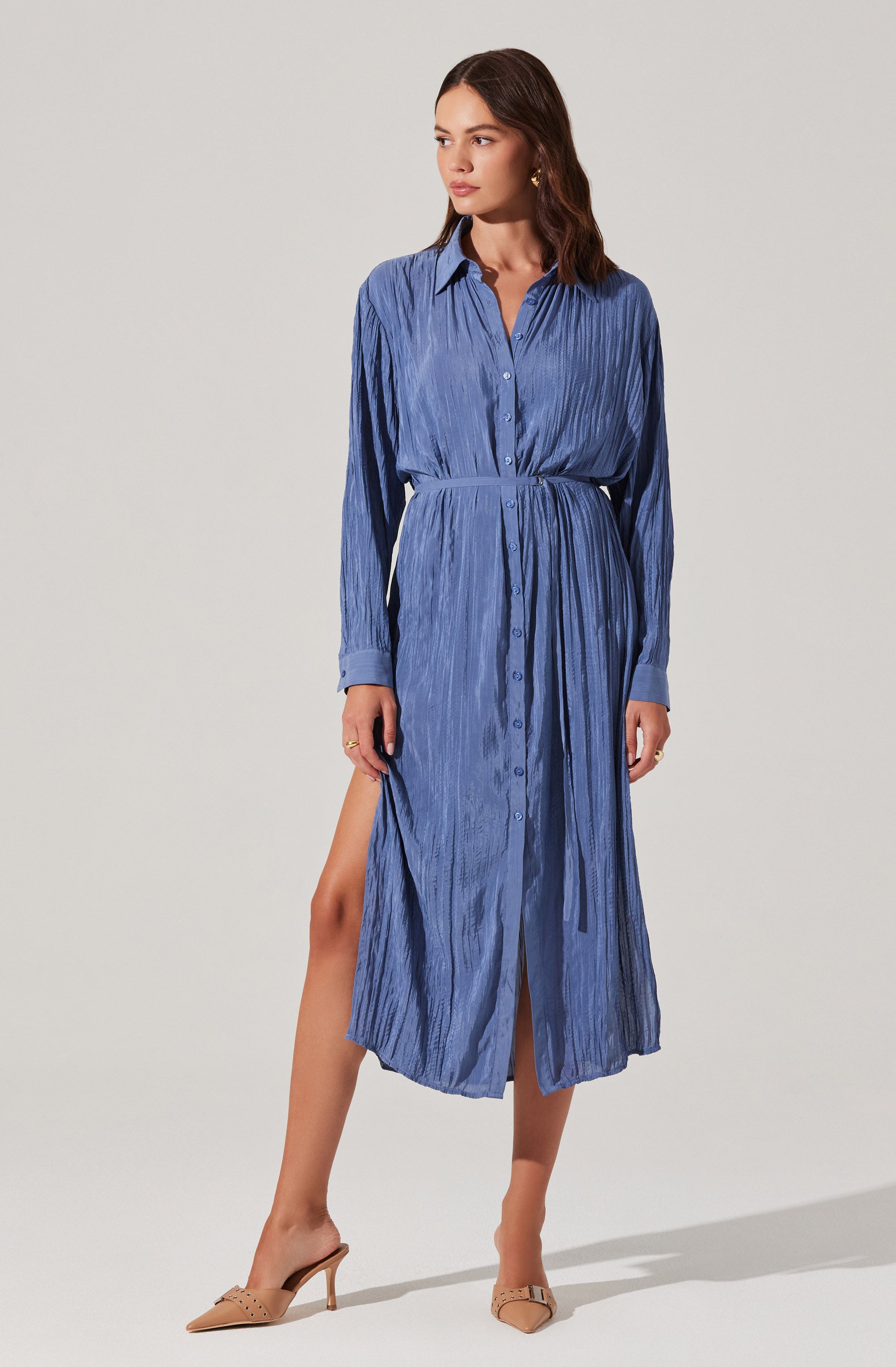 A woman is wearing a long-sleeved, button-down shirt dress in a blue, textured fabric. The dress features a collar, a tied waist, and a slit that reveals one leg. She has paired the outfit with beige pointed-toe high heels.