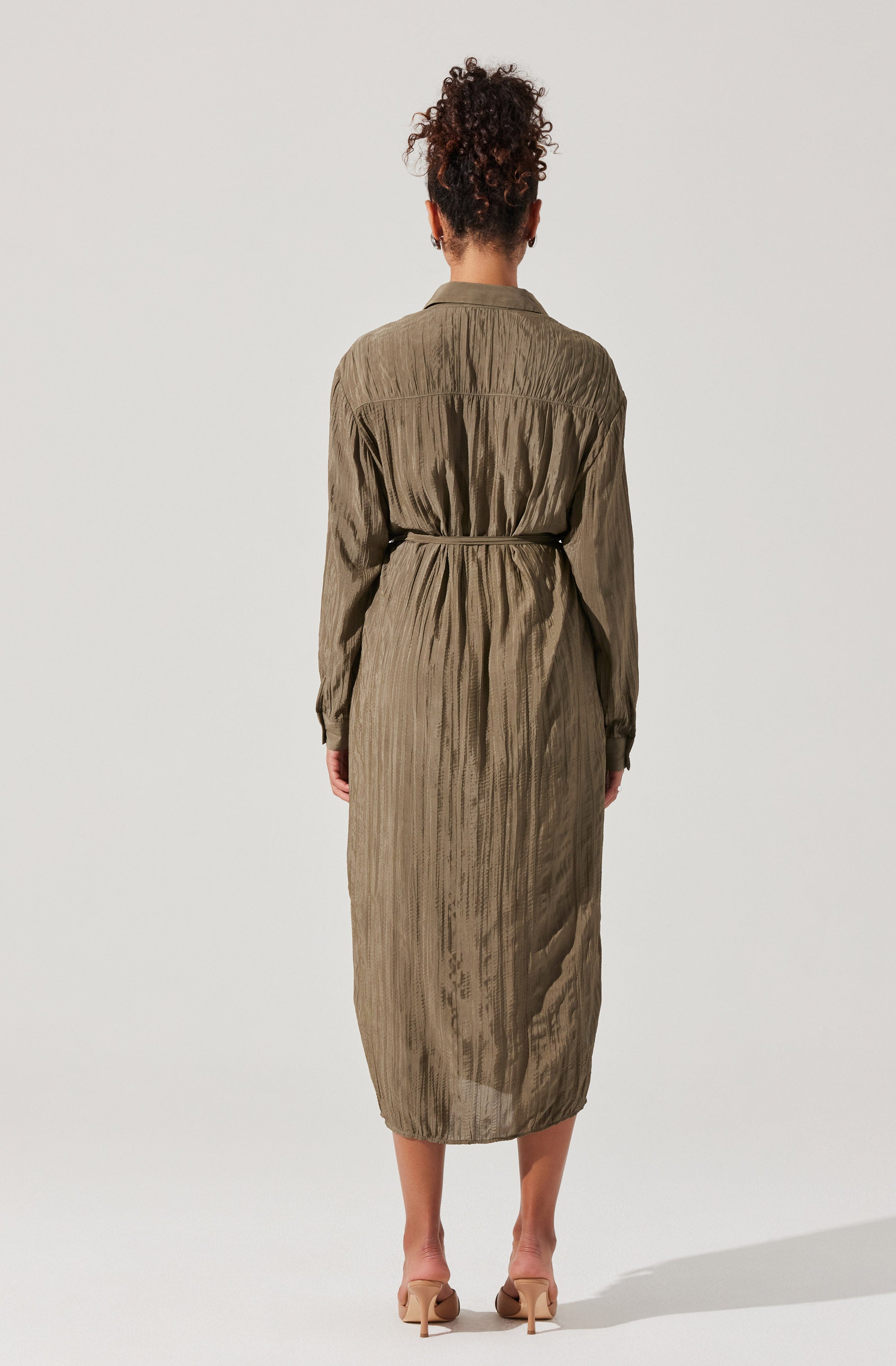 The woman is wearing a long, olive-green dress with a subtle vertical stripe pattern. The dress has long sleeves, a collar, and a waist belt made of the same fabric, which cinches the waist. The material appears lightweight and slightly textured. She is also wearing nude-colored high-heeled shoes.