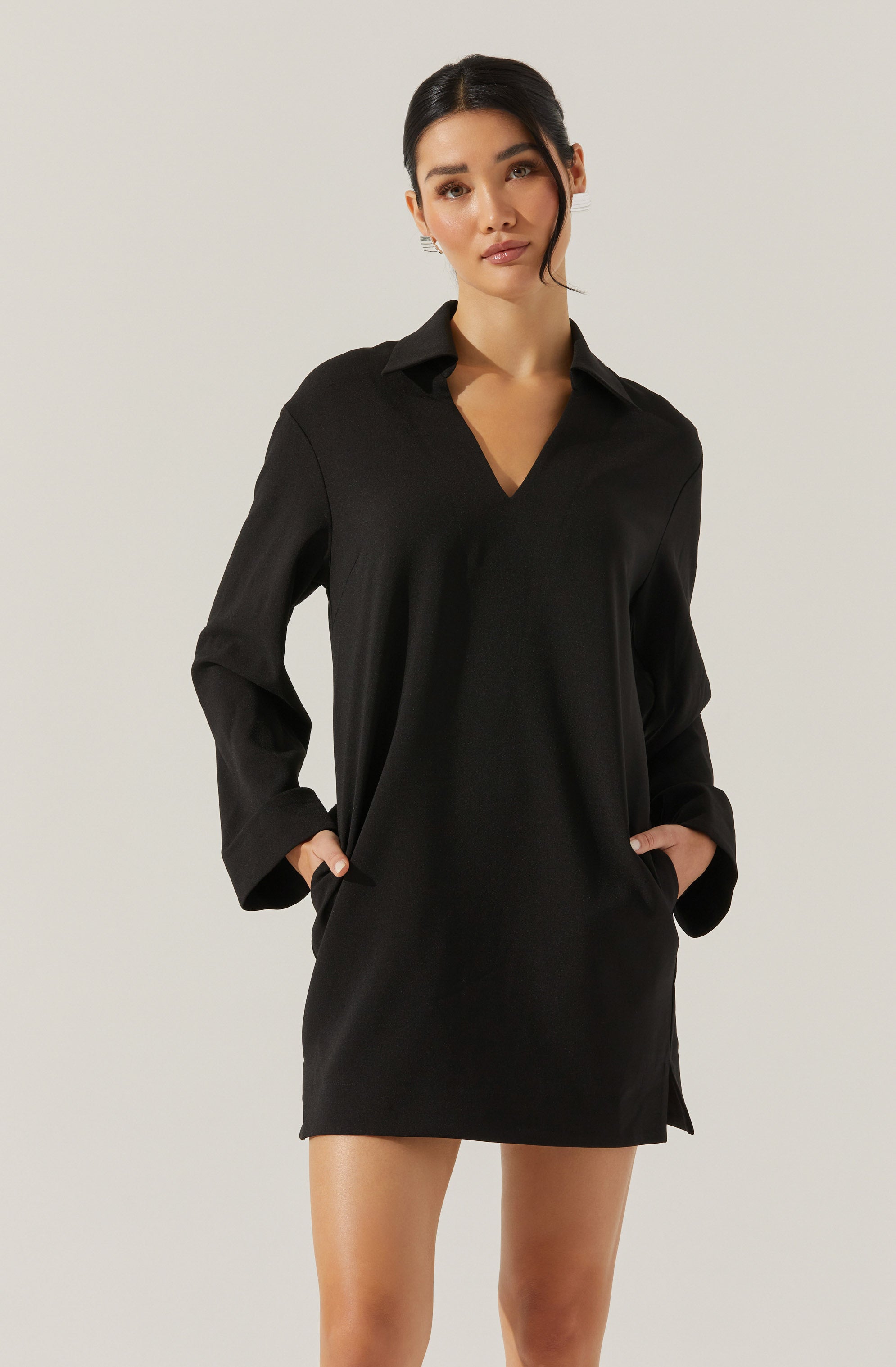 A woman is wearing a black long-sleeve shirt dress with a V-neck collar. The dress has a loose fit, with the sleeves slightly rolled up and side slits at the hem. It appears made of a smooth, lightweight fabric.