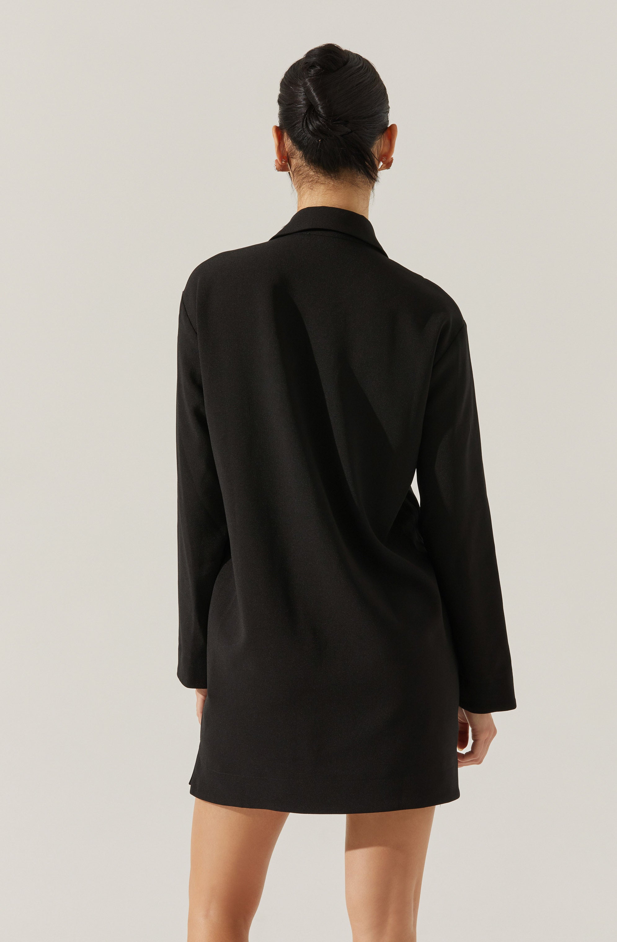 A person wearing a black blazer-style dress with long sleeves. The dress has a tailored fit with a notched lapel collar, and the fabric appears smooth and slightly shiny. The person has dark hair styled in an elegant updo.