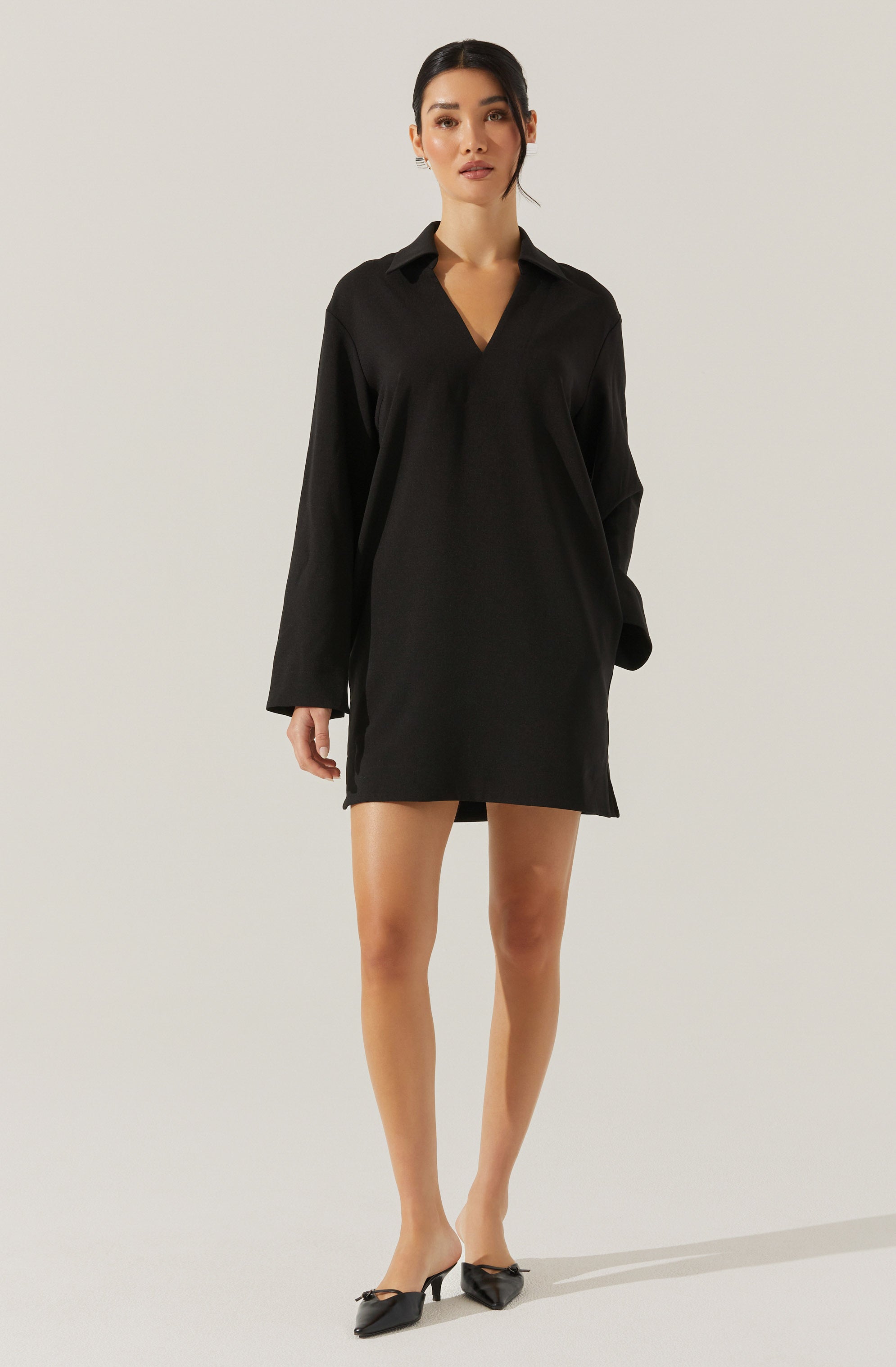 A woman is wearing a loose-fitting, long-sleeved black shirt dress with a V-neck collar. The dress has a simple, minimalist design and falls above the knees. She is also wearing black pointed-toe heels with bows on the front.