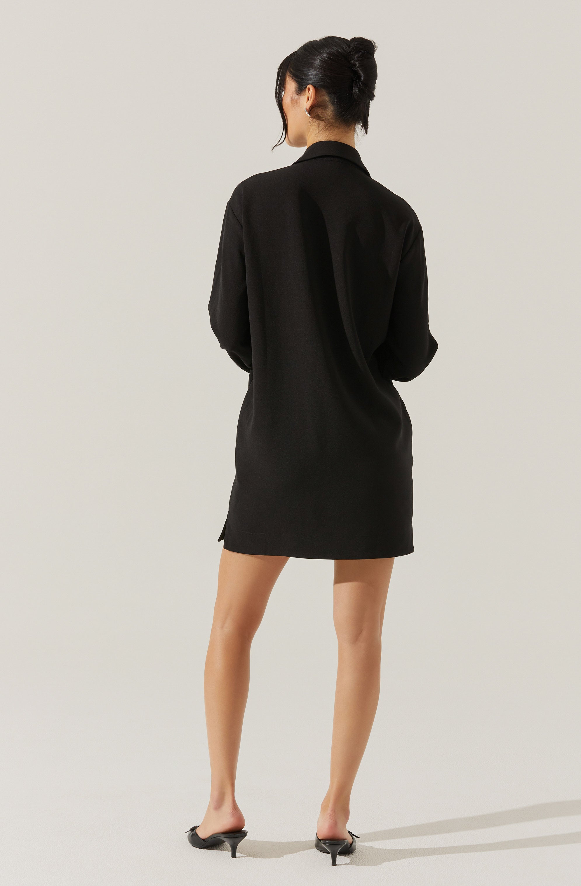 A woman is wearing a black, long-sleeved, collared shirt dress that reaches mid-thigh. The dress is loose-fitting, and she is also wearing black high-heeled shoes with an open toe design. Her hair is styled in an updo, and she is facing away from the camera, standing against a plain light-colored background.