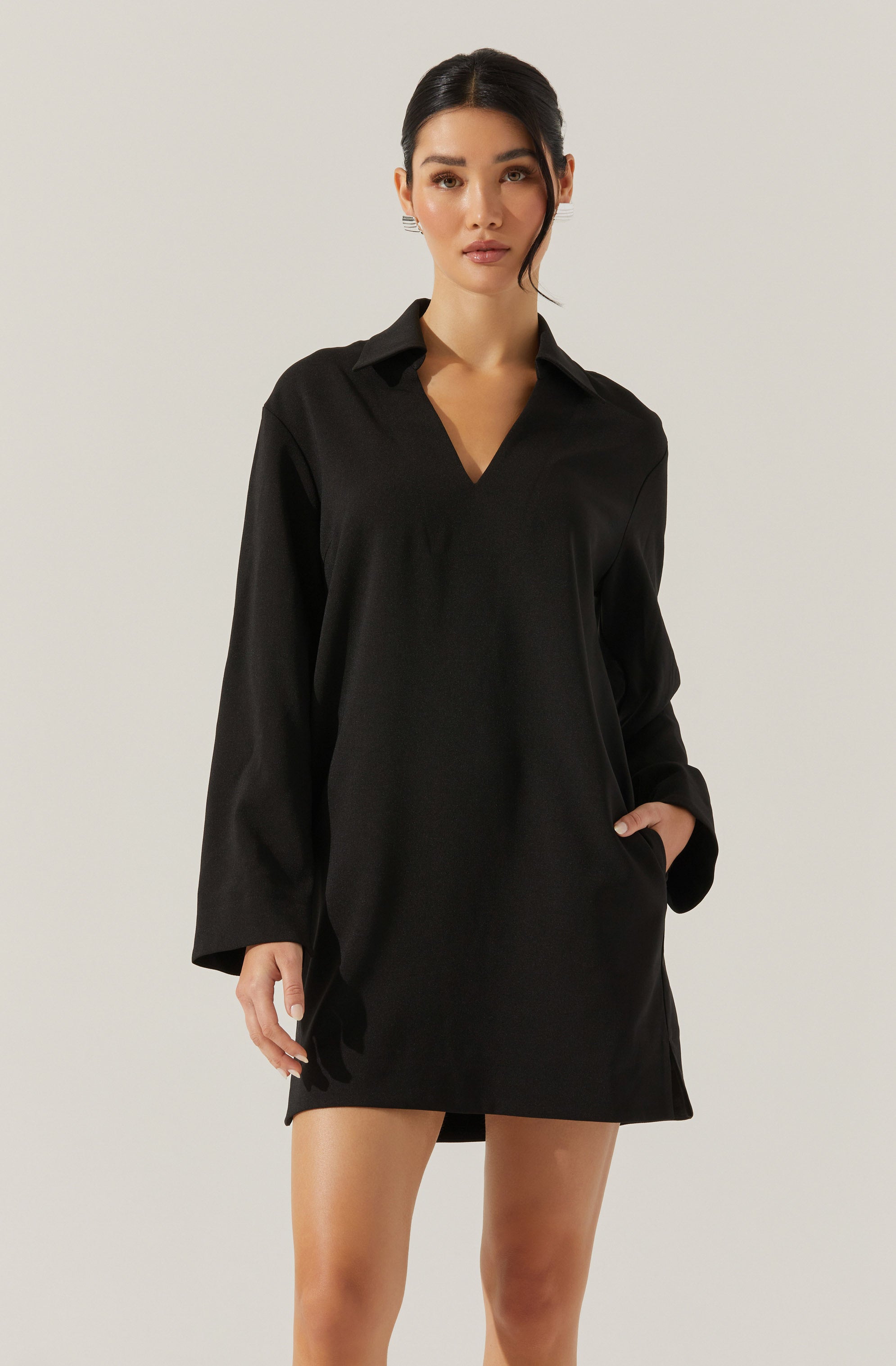 A woman is wearing a loose-fitting, long-sleeved black dress with a collar and a V-neckline. The dress falls above the knees and has a pocket on the right side, where she has resting her hand. The fabric appears smooth and slightly shiny.