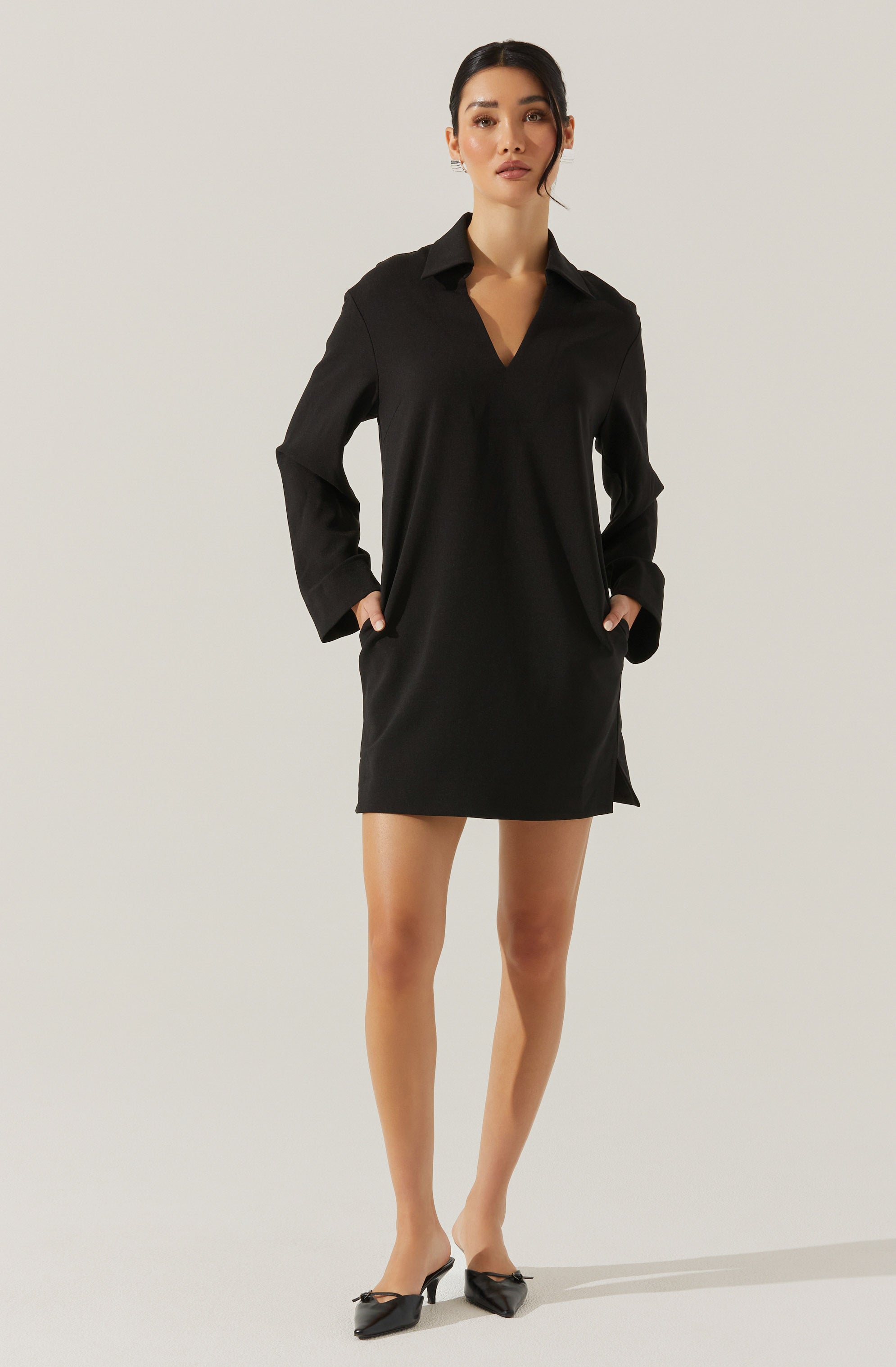 A woman is wearing a long-sleeved black shirt dress with a collar, a V-neckline, and a straight, loose fit that falls above the knees. She has her hands in the dress's side pockets and is wearing black pointed-toe heels with a small bow detail.