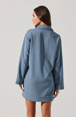 A woman is wearing a light blue, loose-fitting shirt dress with long sleeves and a collar. The dress has a simple, minimal design with no visible embellishments.