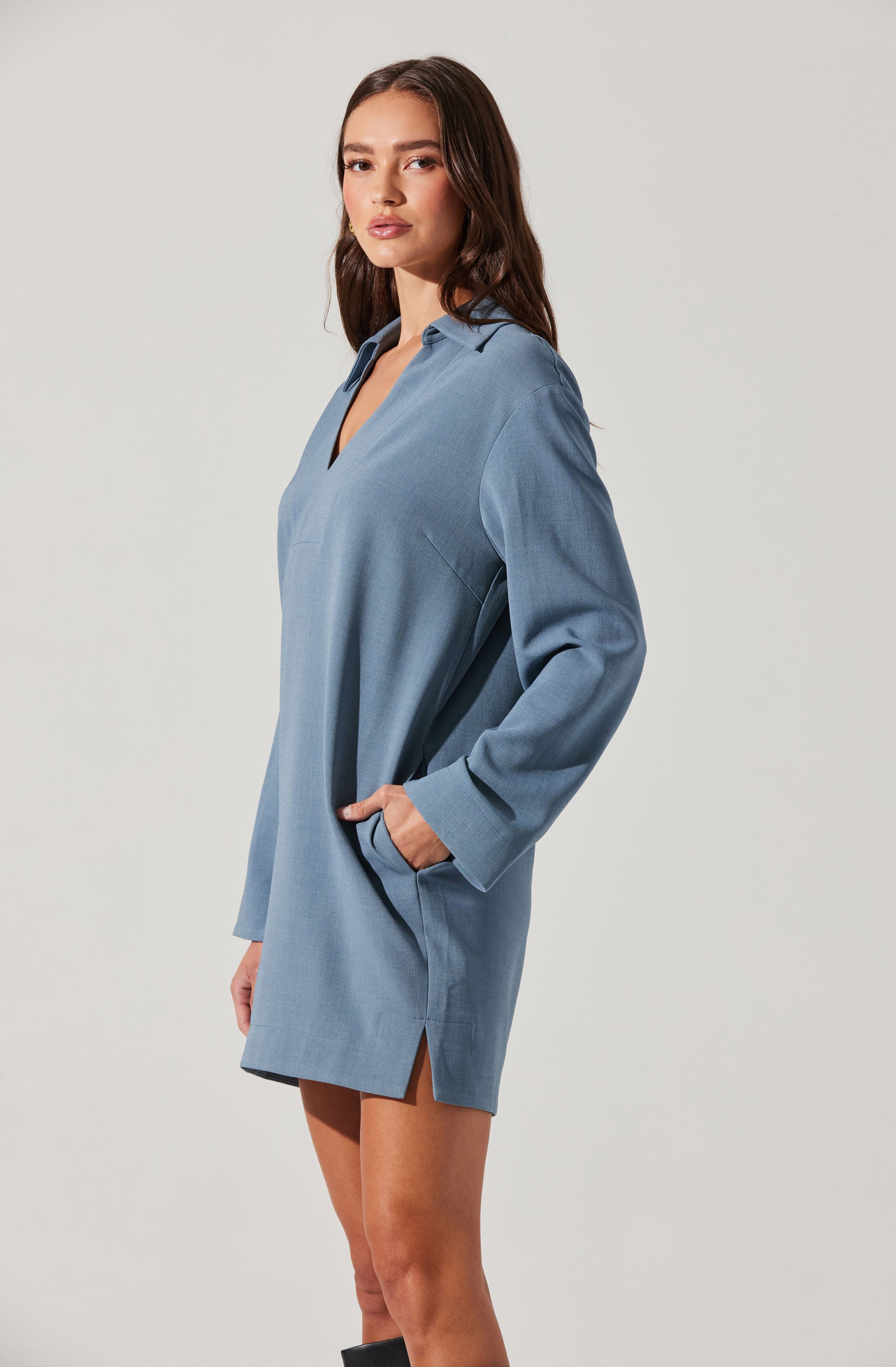 A woman is wearing a light blue, long-sleeved shirt dress. The dress has a collar, a V-neckline, and side pockets. It features a subtle slit on the hem and appears to be made of a soft, lightweight fabric.