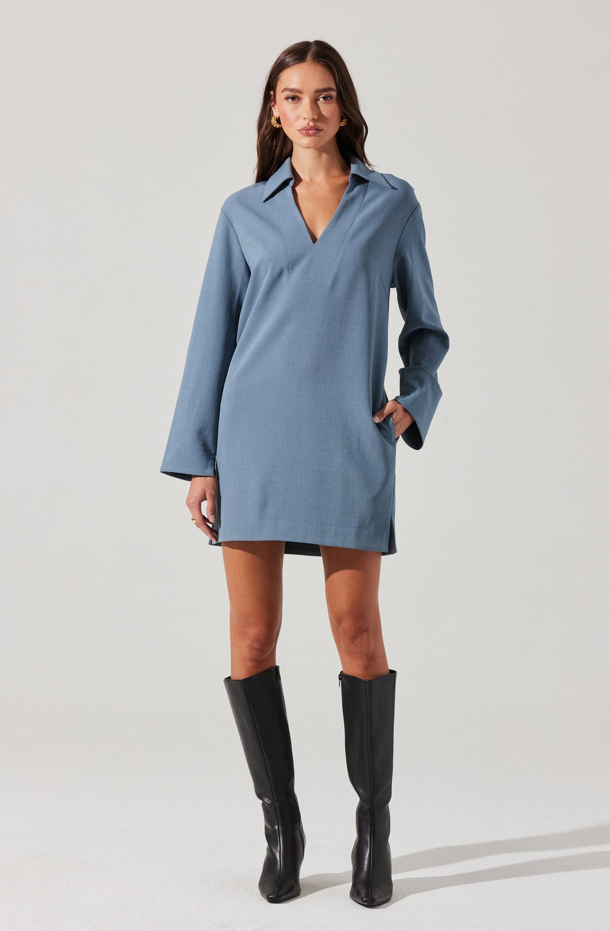 A young woman is wearing a loose-fitting, long-sleeved, light blue shirt dress with a collar and V-neck opening. The dress has a relaxed silhouette that falls above the knees. She pairs the outfit with tall black knee-high boots.
