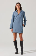 A young woman is wearing a loose-fitting, long-sleeved, light blue shirt dress with a collar and V-neck opening. The dress has a relaxed silhouette that falls above the knees. She pairs the outfit with tall black knee-high boots.