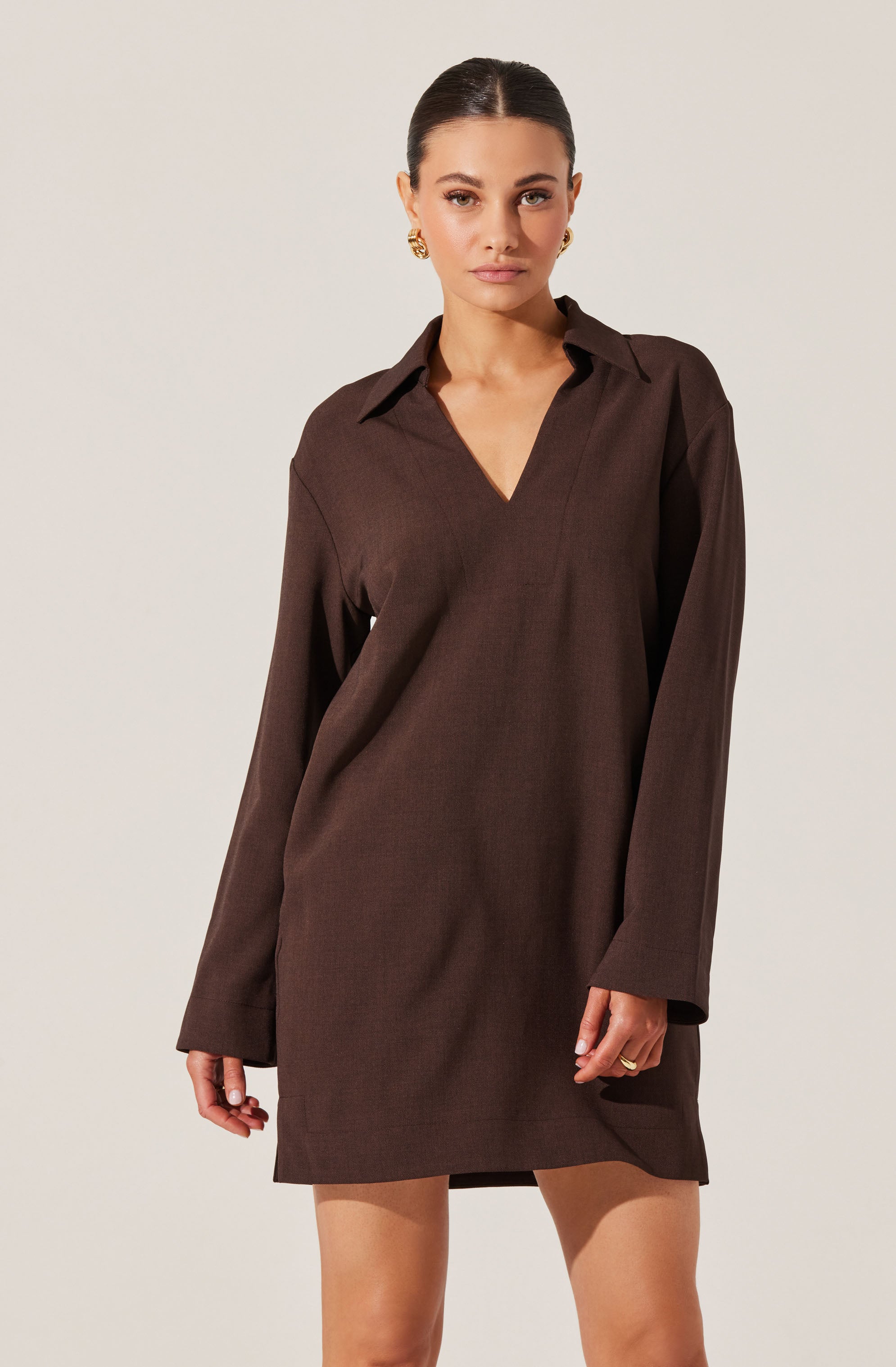 A woman is wearing a loose-fitting, long-sleeved, dark brown shirt dress with a collar and a V-neckline. The dress has a simple, minimal design with no visible patterns or embellishments. She accessorized with gold earrings and rings.