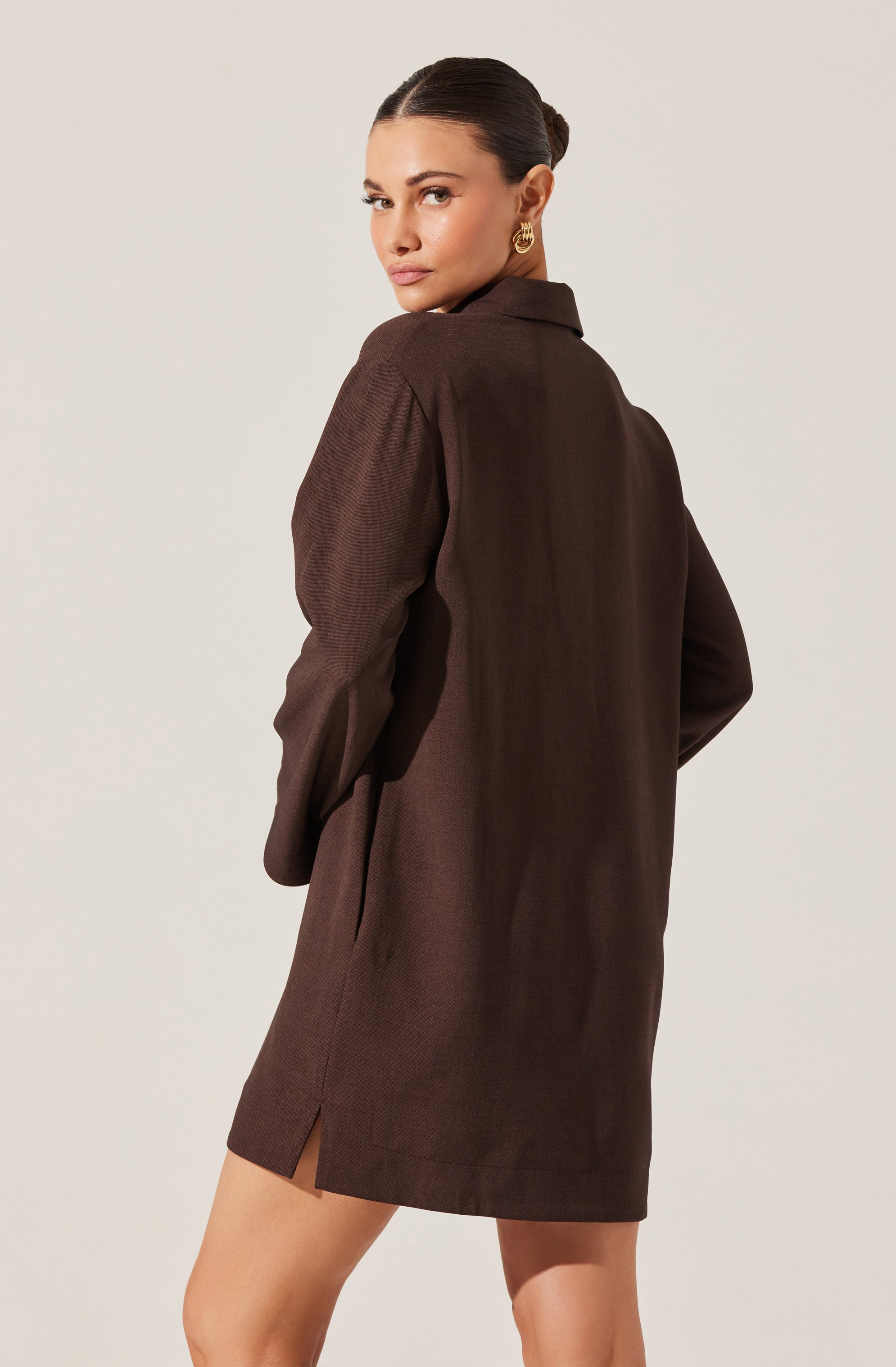 A woman is wearing a long, dark brown shirt dress with long sleeves. The dress has a collar and a slit at the bottom hem on the side. She has her hair styled in a sleek bun and is accessorized with gold hoop earrings. She is standing against a plain, light-colored background, looking over her shoulder.