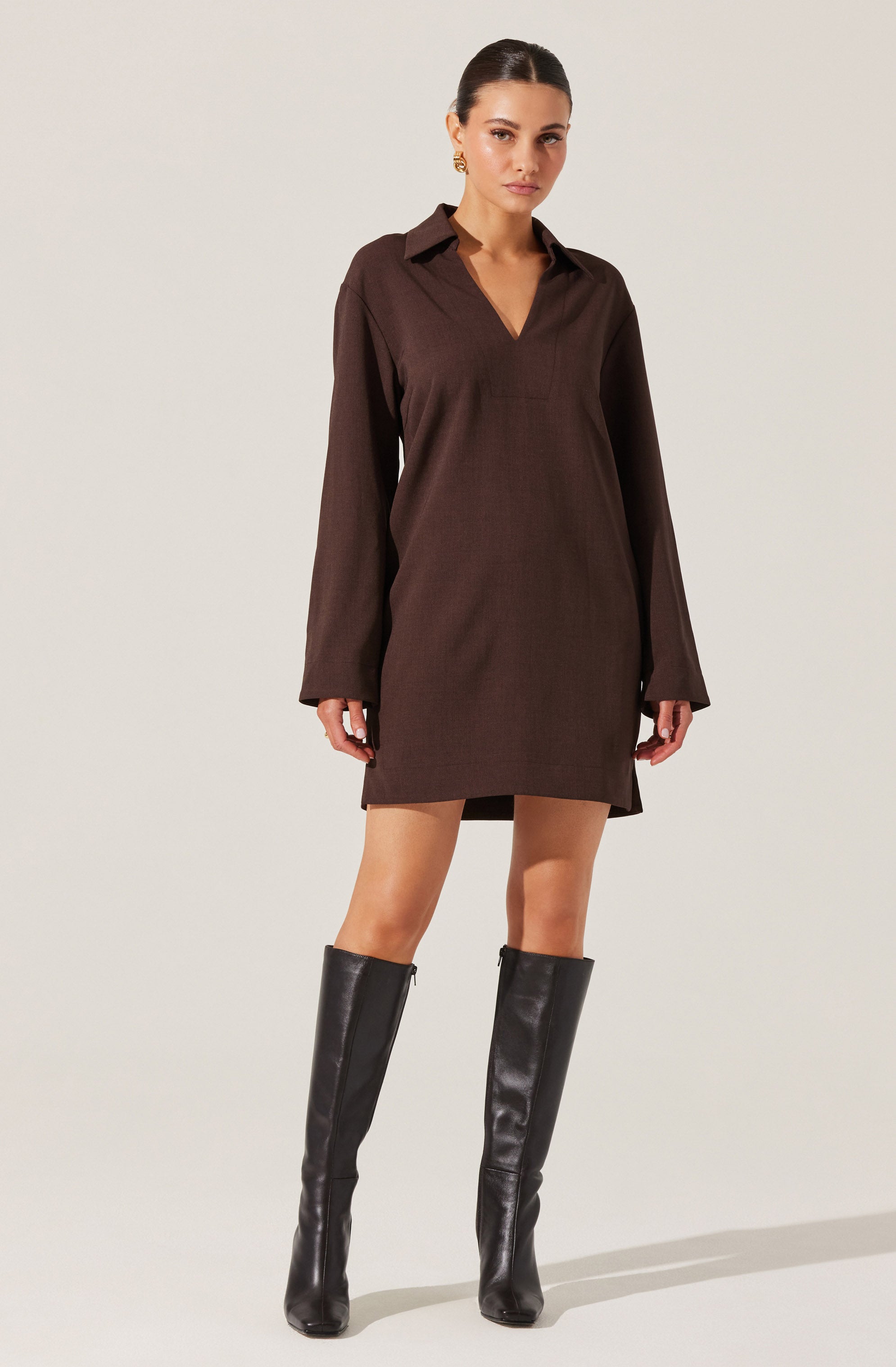 A woman is wearing a long-sleeved, dark brown shirt dress with a collar and a V-neckline. The dress is loose-fitting and falls above the knees. She is also wearing tall, black leather knee-high boots.
