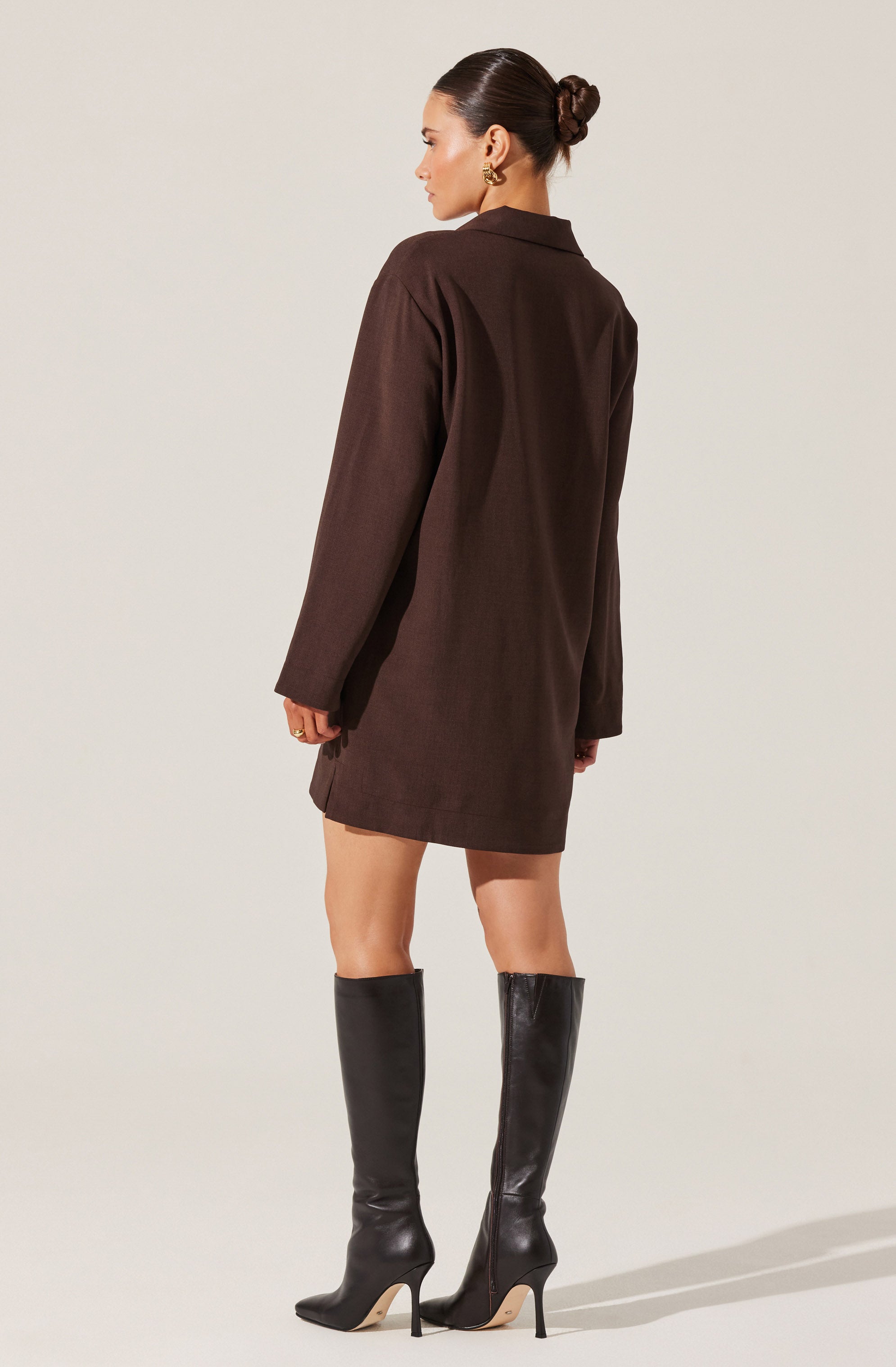 A woman is wearing a brown oversized blazer that extends to mid-thigh, with long sleeves and a collar. She has paired it with tall black leather high-heeled boots that reach just below the knees.
