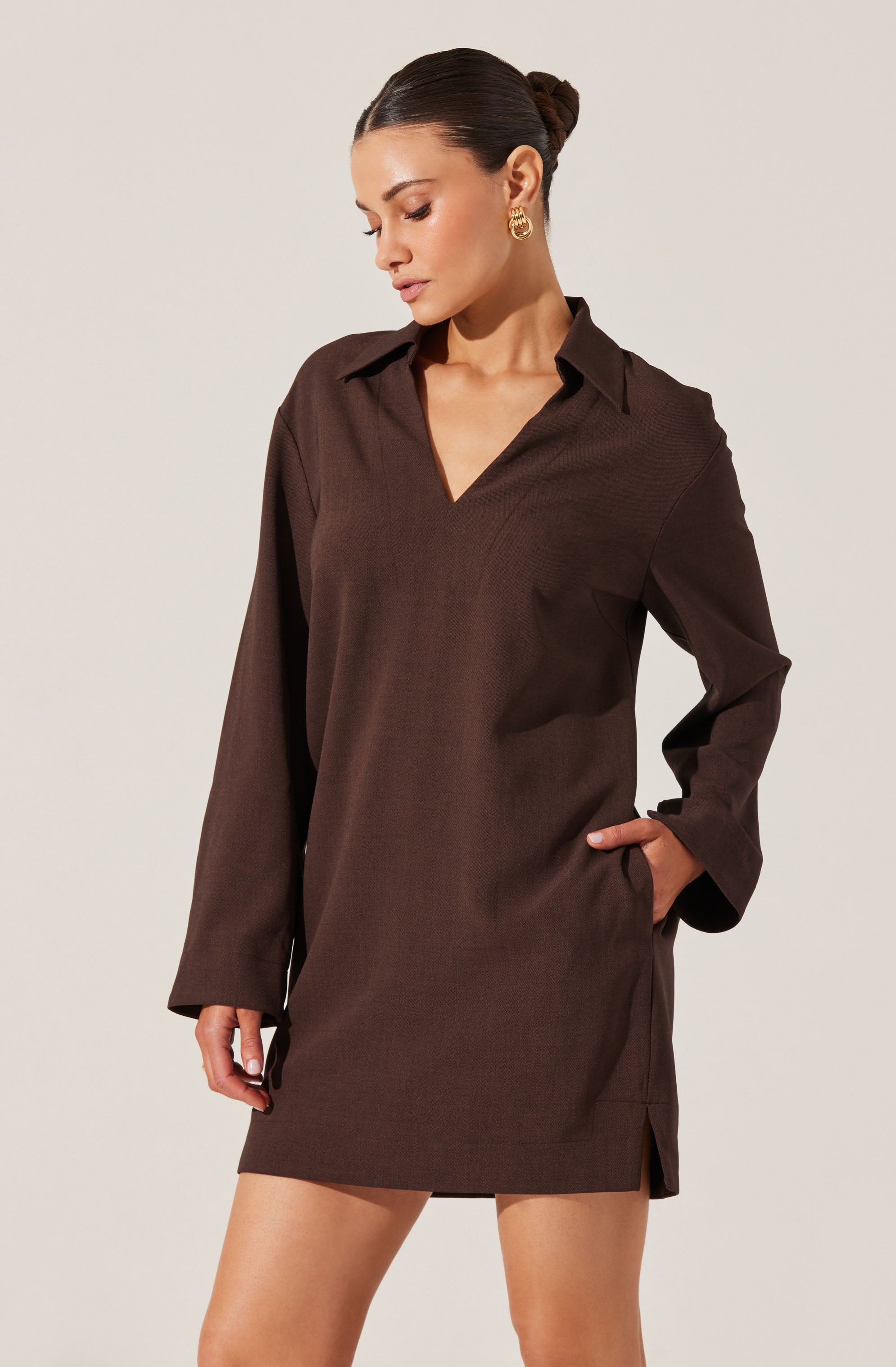 A woman is wearing a brown, long-sleeved shirt dress with a collar and V-neckline. The dress is loose-fitting, with the hem ending above the knees and featuring small side slits. She has her left hand in her pocket and is wearing gold hoop earrings. Her hair is styled in a sleek, low bun.