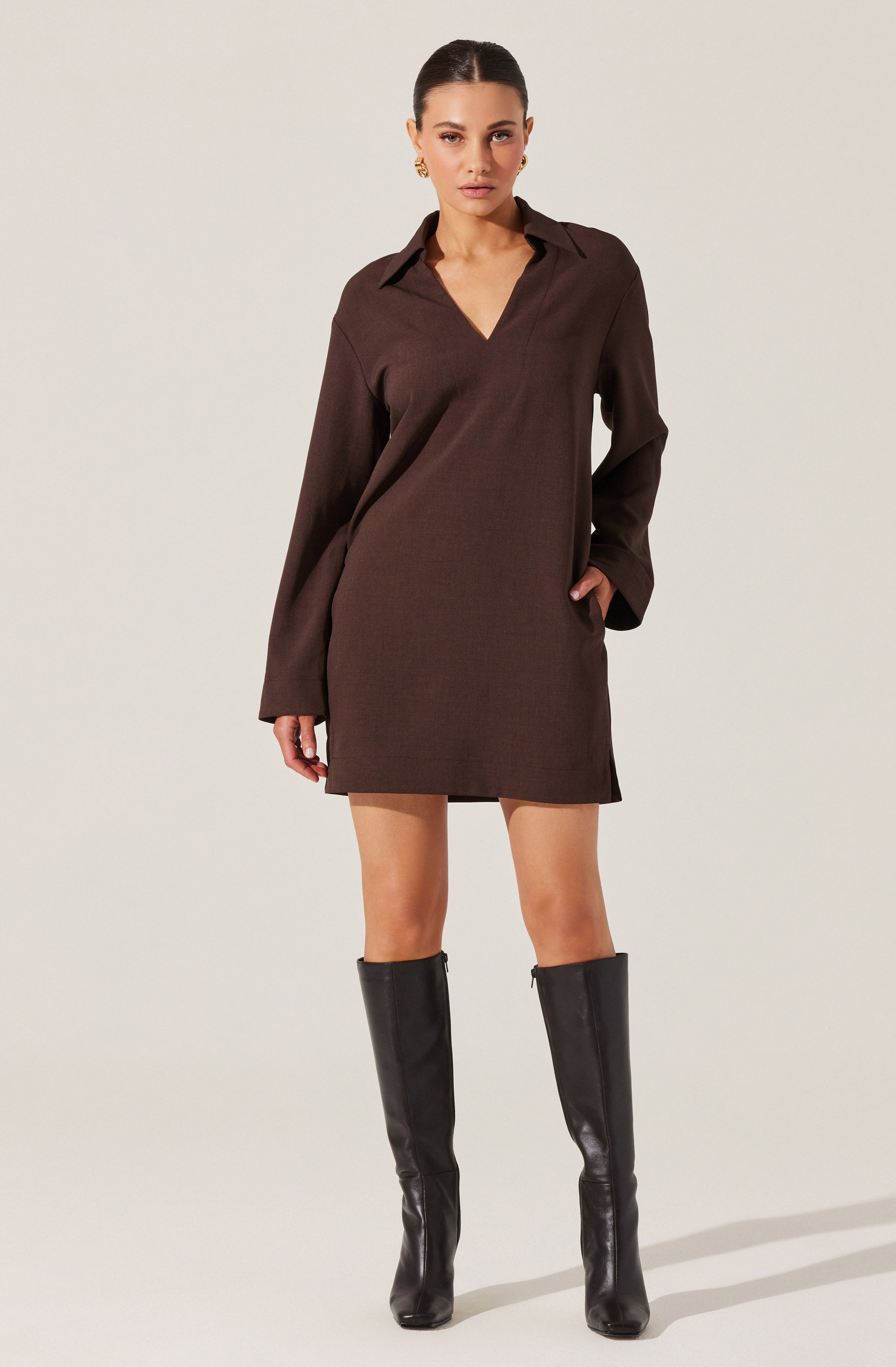 A woman is wearing a loose-fitting, long-sleeved brown dress with a collar and V-neckline. The dress falls above the knees. She is also wearing tall black leather boots that reach just below her knees.
