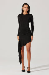 A woman is wearing a fitted, long-sleeved black dress with a high neckline. The dress features a unique asymmetrical hemline, with a ruffled detail cascading from one side, shorter in the front and longer in the back. She pairs the outfit with black ankle-strap high heels.