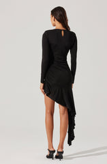 A woman is wearing a fitted black long-sleeve top with a small keyhole opening at the back neckline. She is also wearing a high-low black skirt with a ruffled, asymmetrical hem that drapes down to her ankle on one side. She has on black high-heeled sandals.