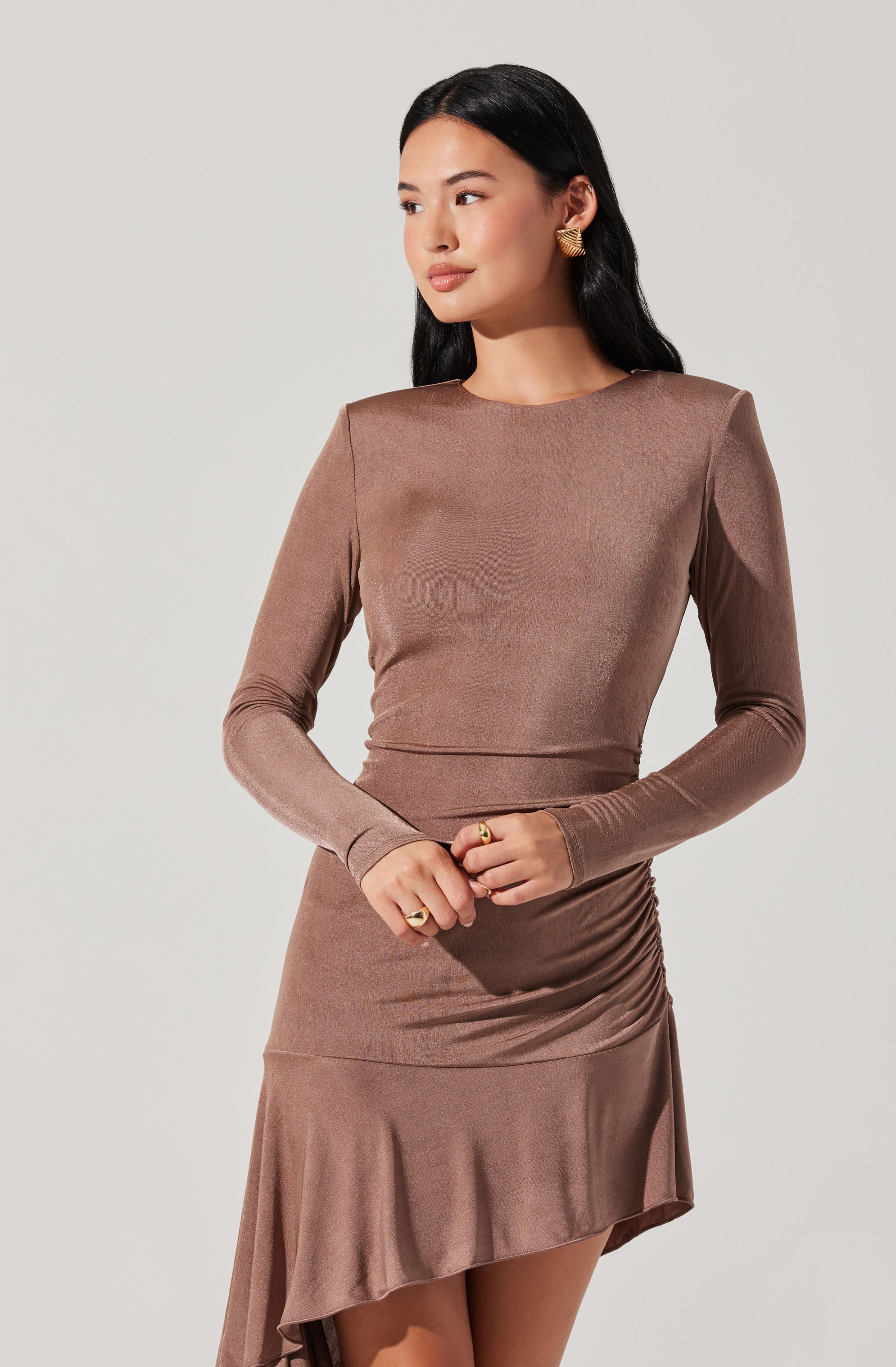 A woman wearing a fitted, long-sleeved, taupe-colored dress with a round neckline. The dress features a ruched side detail on the skirt and has a slight asymmetrical hem with a ruffled edge. She has long, dark hair and is accessorized with gold earrings and rings.