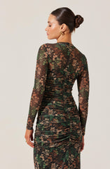 A woman is wearing a fitted, long-sleeved dress with a floral pattern. The dress features a mix of earthy tones, including green, brown, and beige, and has a ruched design along the back, accentuating her figure. She has her dark hair styled in a sleek bun and is accessorized with gold hoop earrings.