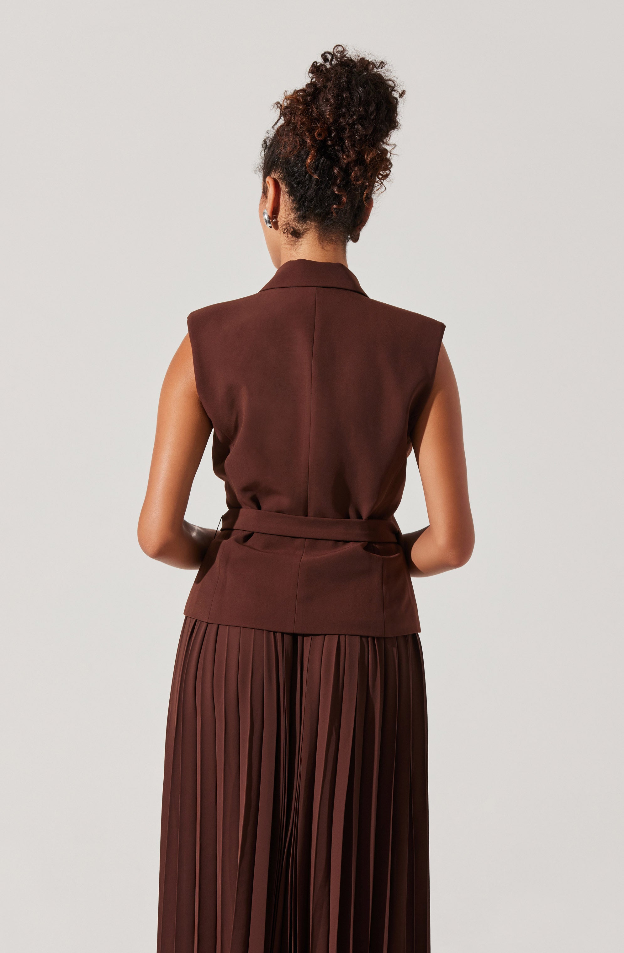 The woman is wearing a sleeveless, brown vest with a collar and a matching waist belt. She is also wearing a long, pleated skirt in a matching brown color.