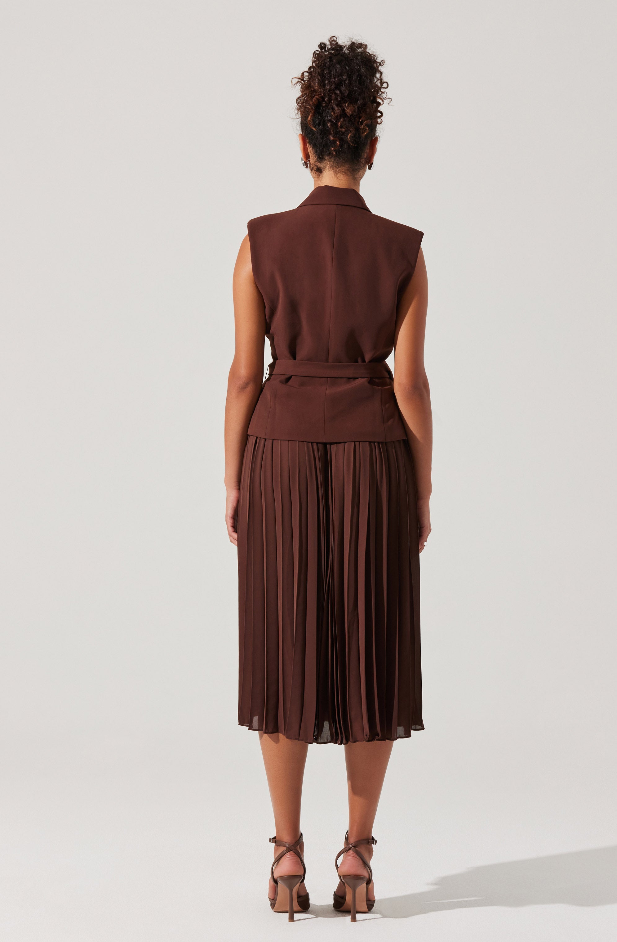 A woman is wearing a sleeveless, tailored brown vest with a belt at the waist, paired with a pleated brown midi skirt that falls below the knees. She is also wearing strappy high-heeled sandals.