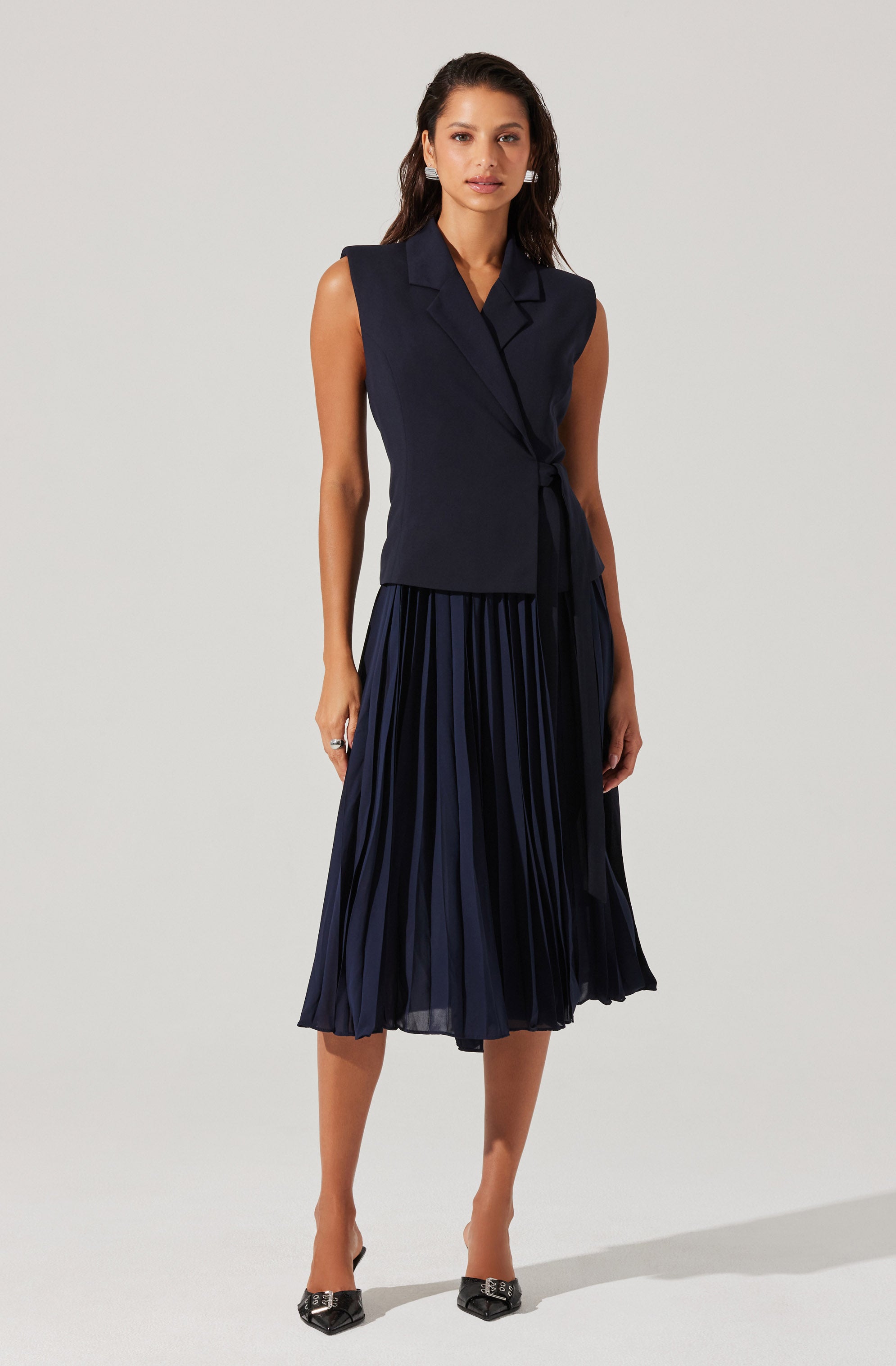 A woman is wearing a sleeveless navy blue vest with a collar and a tie at the waist, paired with a navy blue pleated midi skirt. She has medium-length wavy hair and is standing against a plain light gray background. She is also wearing black pointed-toe shoes with decorative accents.