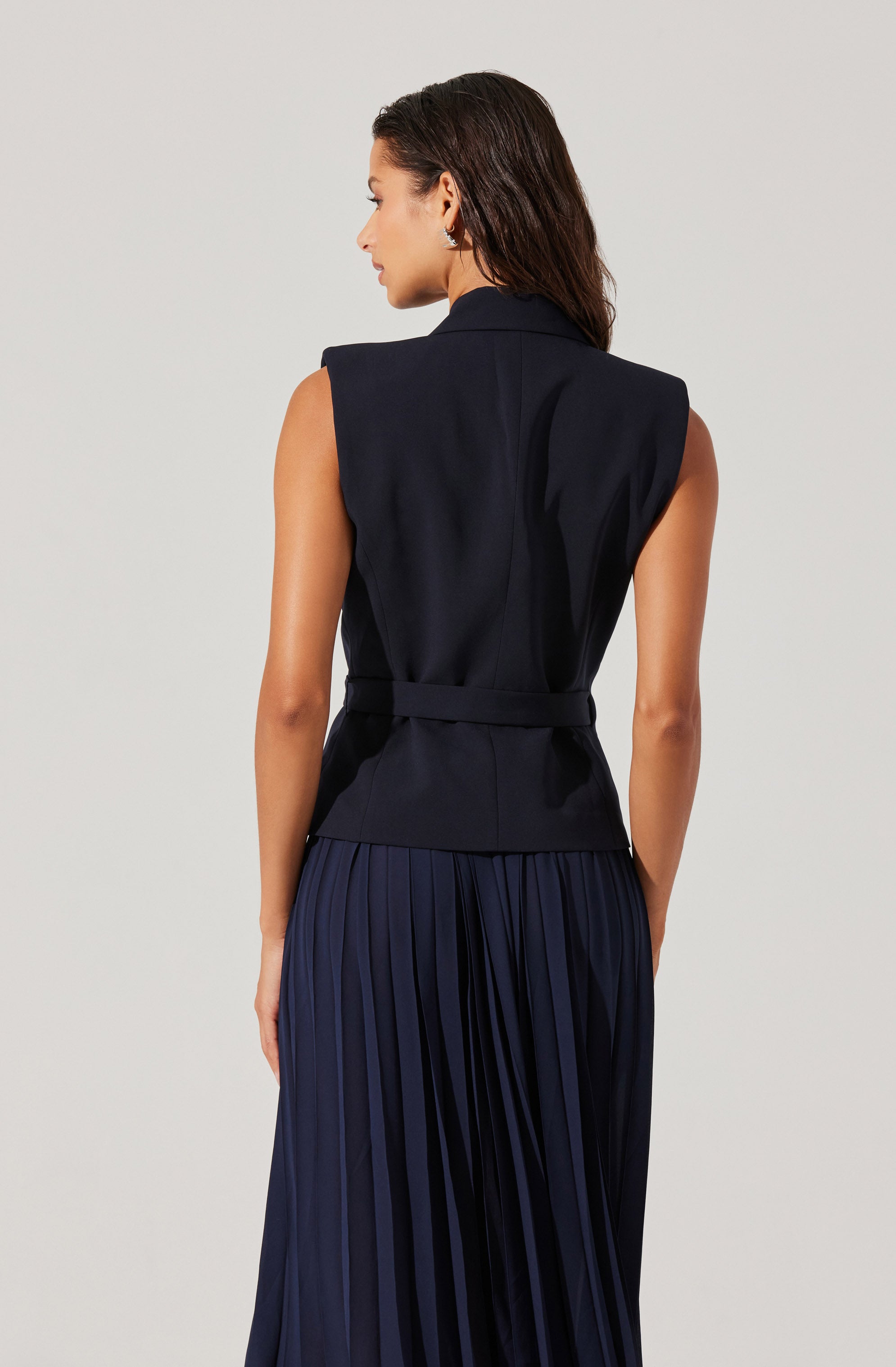 A woman is wearing a sleeveless navy blazer with padded shoulders and a matching fabric belt at the waist. She pairs it with a navy pleated skirt.