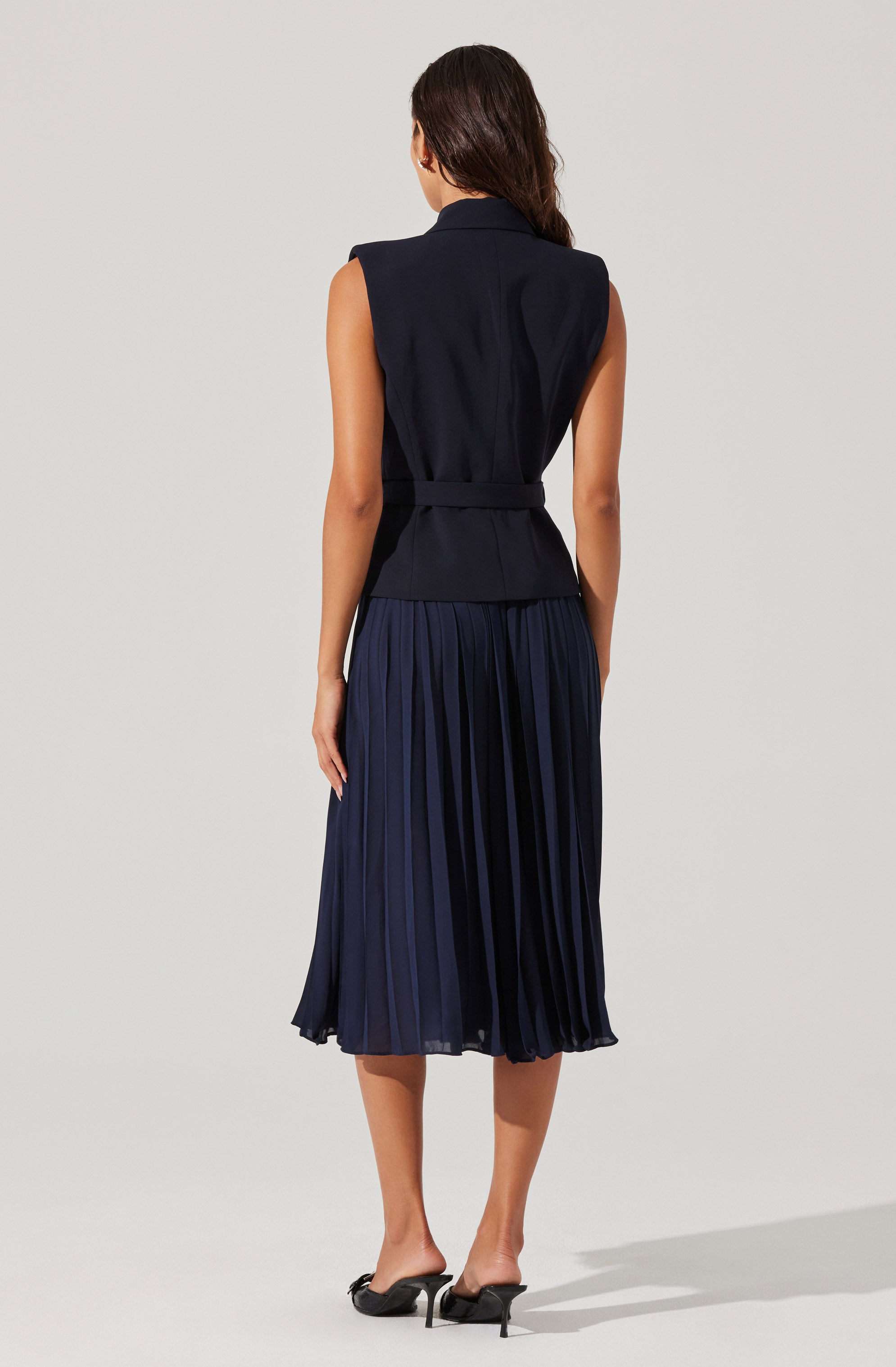 A woman is wearing a sleeveless, dark navy vest with a fitted waist, paired with a matching pleated midi skirt in a similar dark navy color. She also wears black heeled shoes.