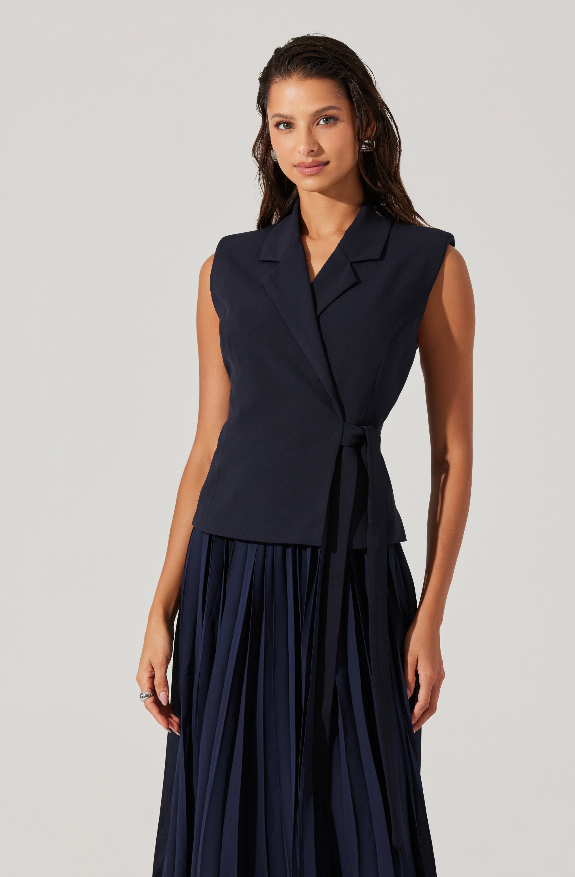 A woman is wearing a sleeveless, dark navy tailored vest with a notched lapel collar and a tie waist. She pairs it with a matching pleated skirt that has vertical stripes, also in dark navy.
