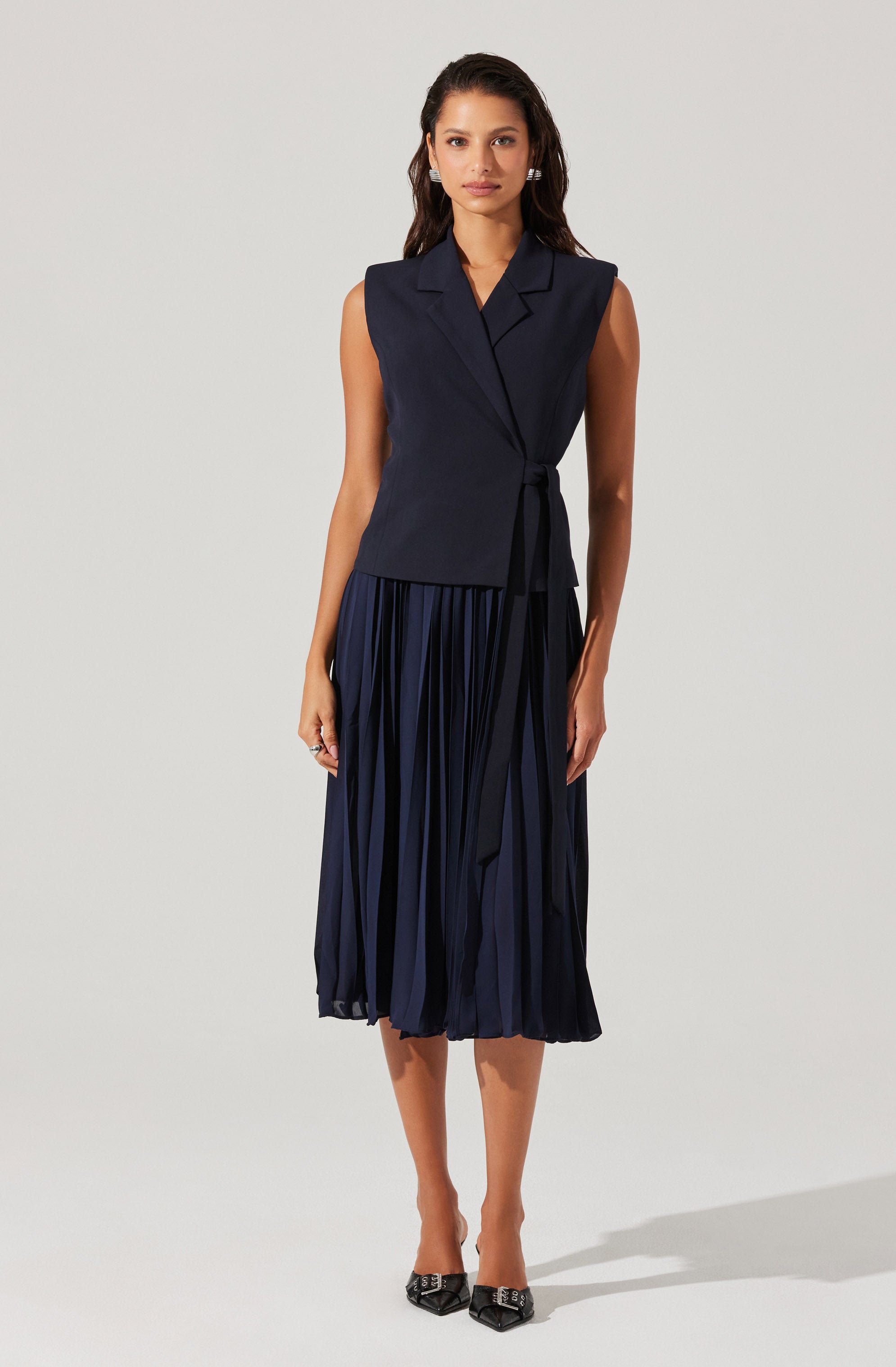 A woman is wearing a sleeveless navy blue vest with a notched lapel collar, tied at the waist with a matching fabric belt. She pairs it with a pleated midi skirt in a similar dark navy color, creating a coordinated outfit. She completes the look with pointed black shoes featuring a decorative buckle.