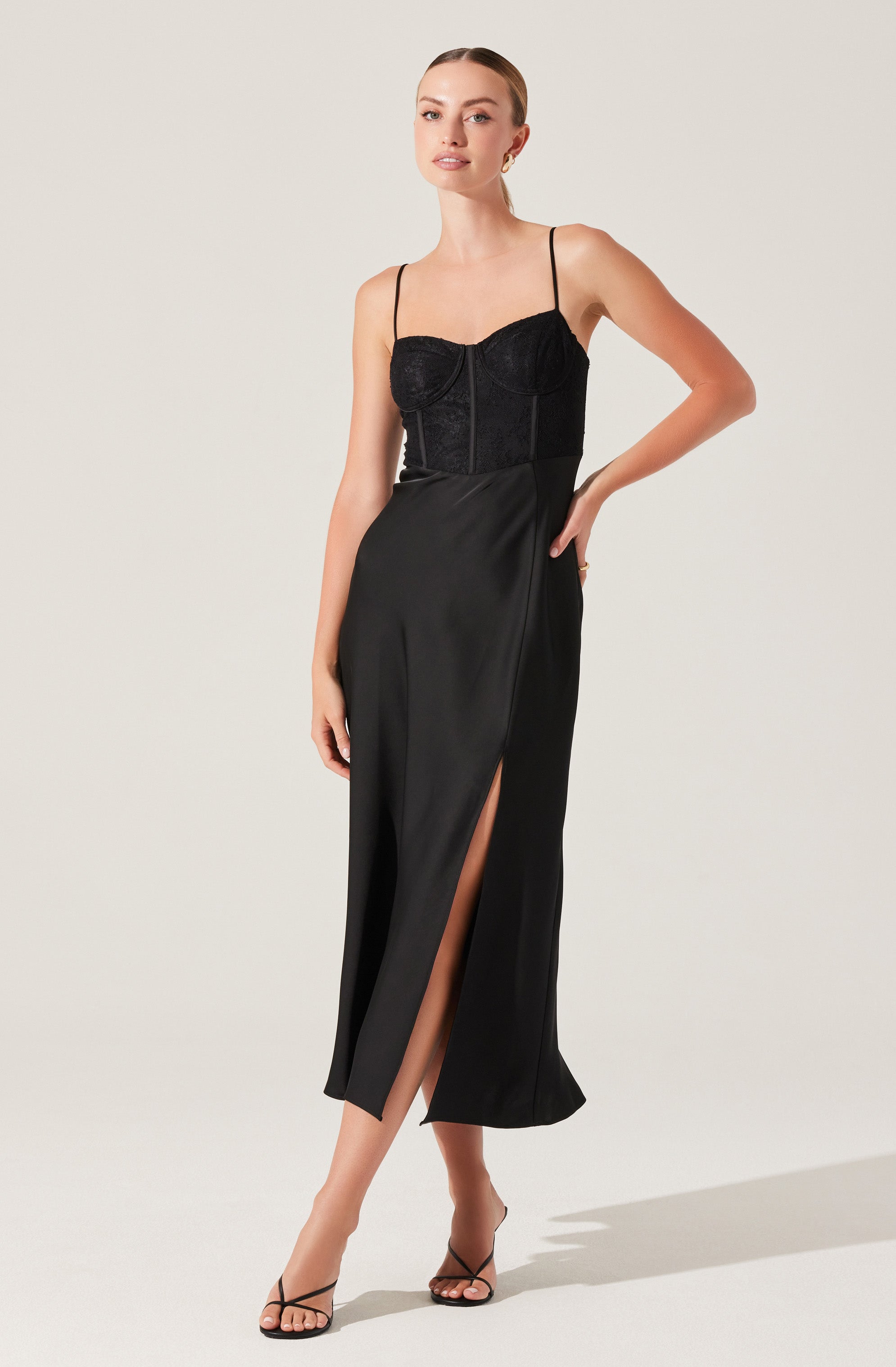 A woman is wearing a black dress with thin spaghetti straps and a fitted bodice made of lace fabric. The dress has a high waistline, with a smooth, flowing skirt that features a front slit revealing her leg. She is also wearing black open-toe high-heeled sandals.