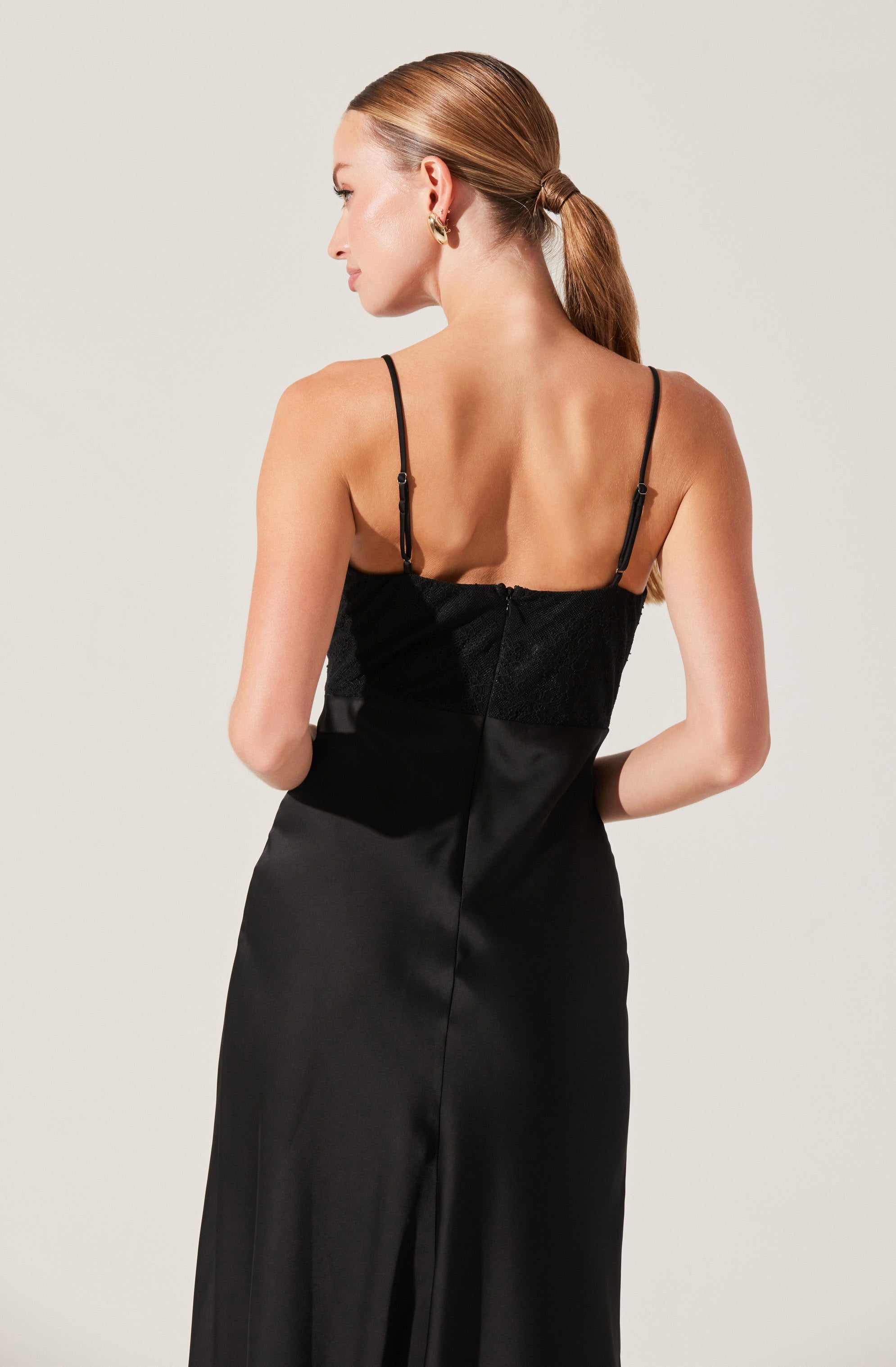 A woman is wearing a black spaghetti strap dress with a fitted bodice made of a lace and mesh material, and a smooth satin-like skirt that extends to the floor.