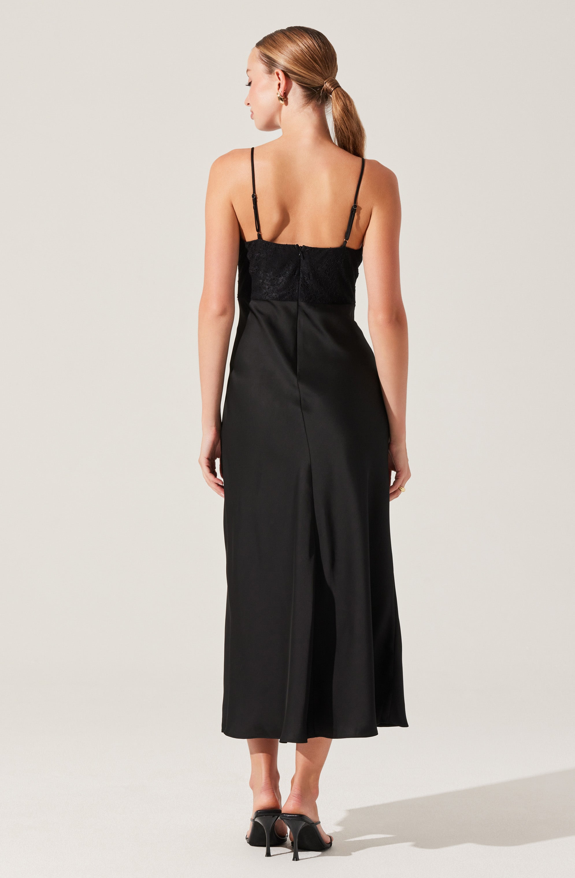 A woman is wearing a black evening dress with thin spaghetti straps. The top part of the dress features a lace or textured fabric, and the skirt is smooth and long, reaching down to her ankles. She is wearing black high-heeled sandals. Her hair is styled in a low ponytail.