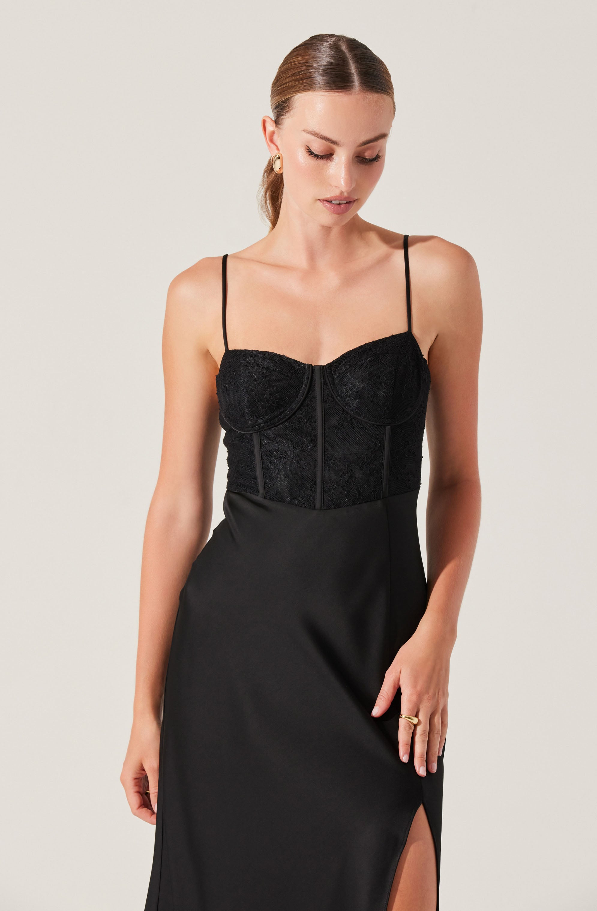 A woman is wearing a black dress with thin spaghetti straps. The bodice of the dress is made of a textured, lace-like fabric with structured cups and vertical seam details. The skirt is smooth, flowing, and features a thigh-high slit on one side.