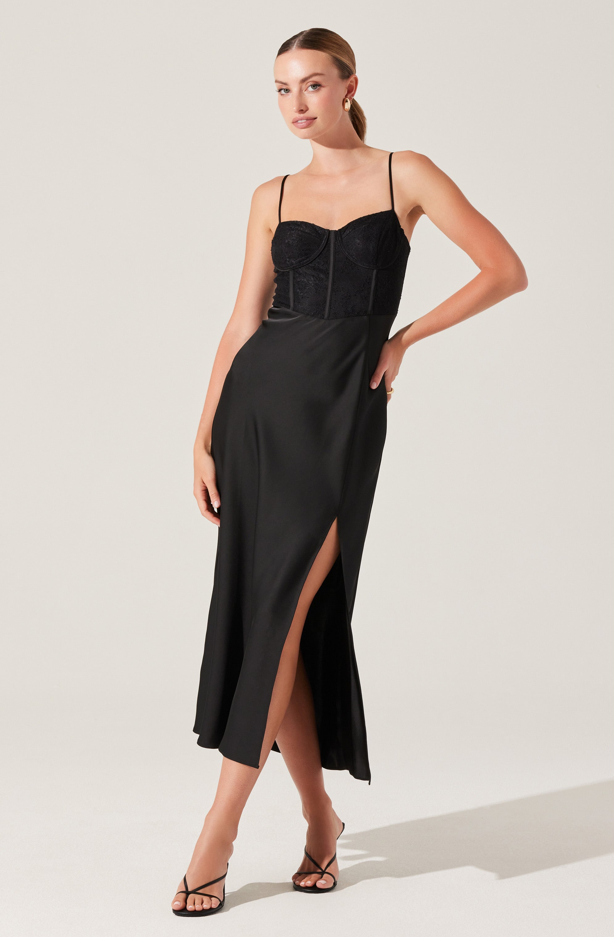 The woman is wearing a black dress with a fitted bodice featuring thin spaghetti straps. The bodice has a zipper detail in the center front. The skirt is long and flowing with a high slit on one side, revealing her leg. She is also wearing black, strappy high-heeled sandals.
