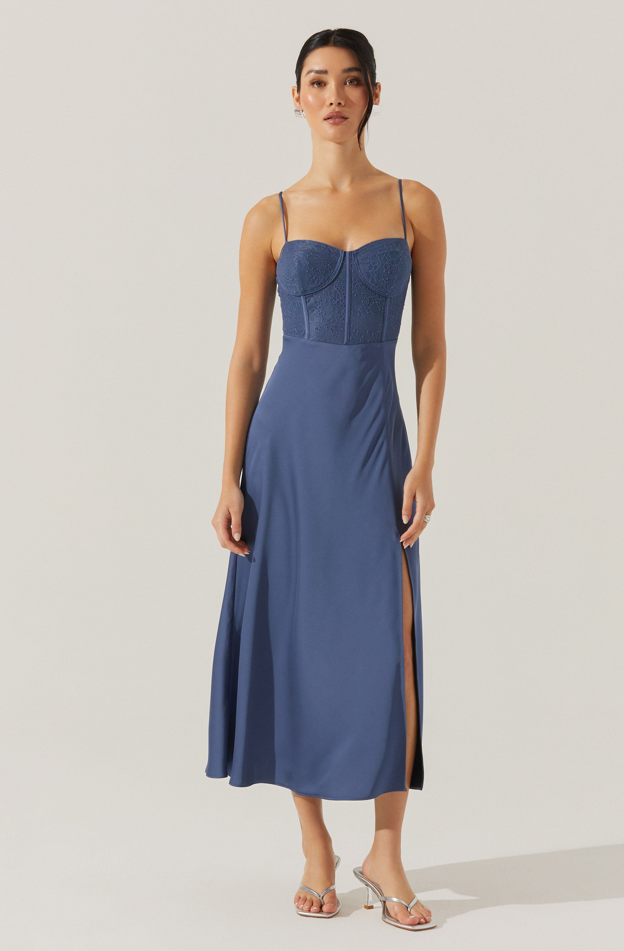A woman is wearing a long, elegant evening dress that combines a fitted, textured, dark blue bodice with thin spaghetti straps. The dress features a flowing skirt in a matching shade of blue with side slits that reveal her legs. She is also wearing a pair of open-toe, clear-strap high-heeled sandals.