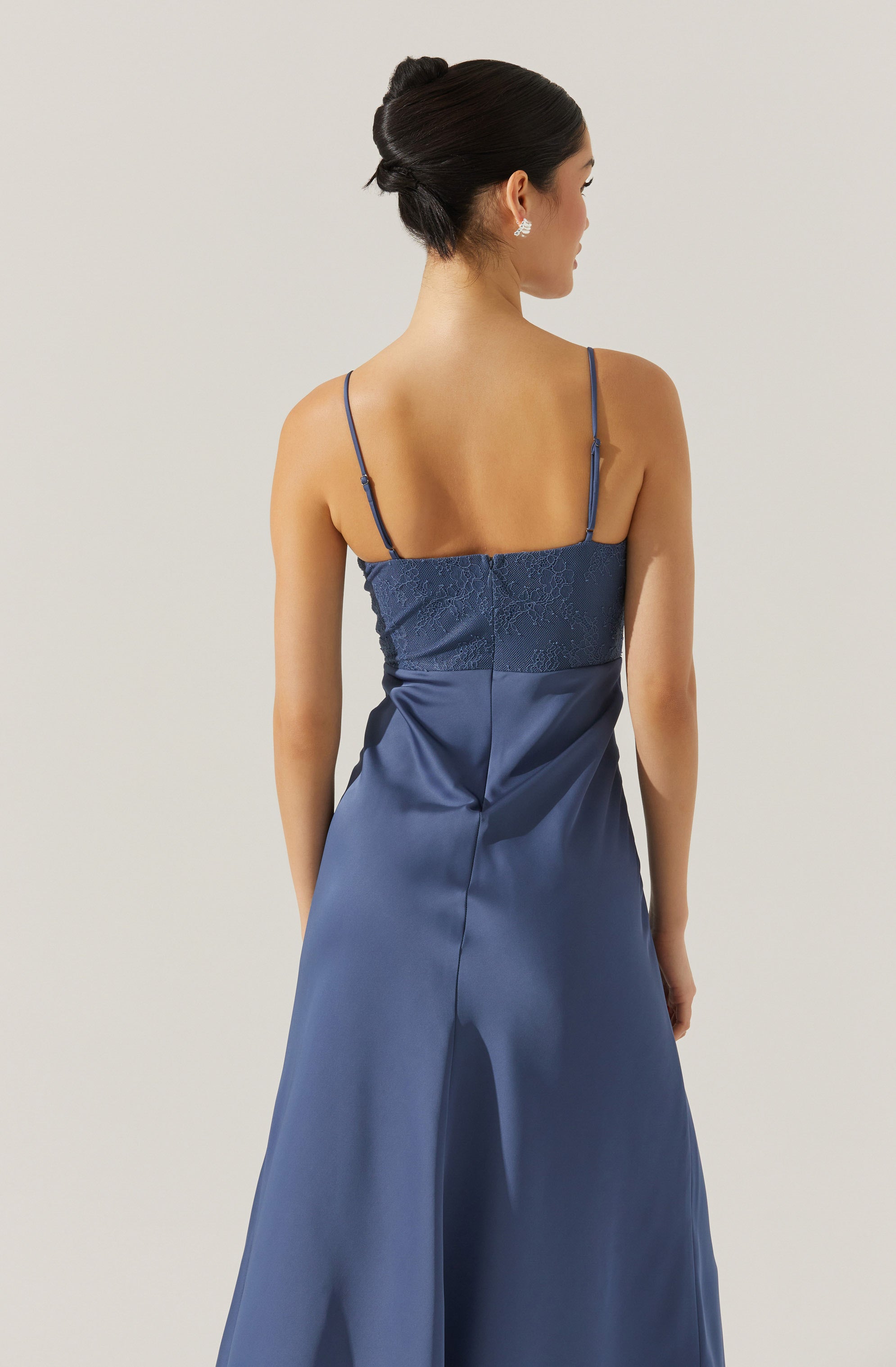 A woman is wearing a navy blue evening gown with thin spaghetti straps. The top part features lace or embroidered detailing, and the fabric of the skirt is shiny and smooth, made from a satin-like material. The dress has a fitted waist and flows flared to the floor. The woman has dark hair styled in an elegant updo and is wearing sparkling earrings.