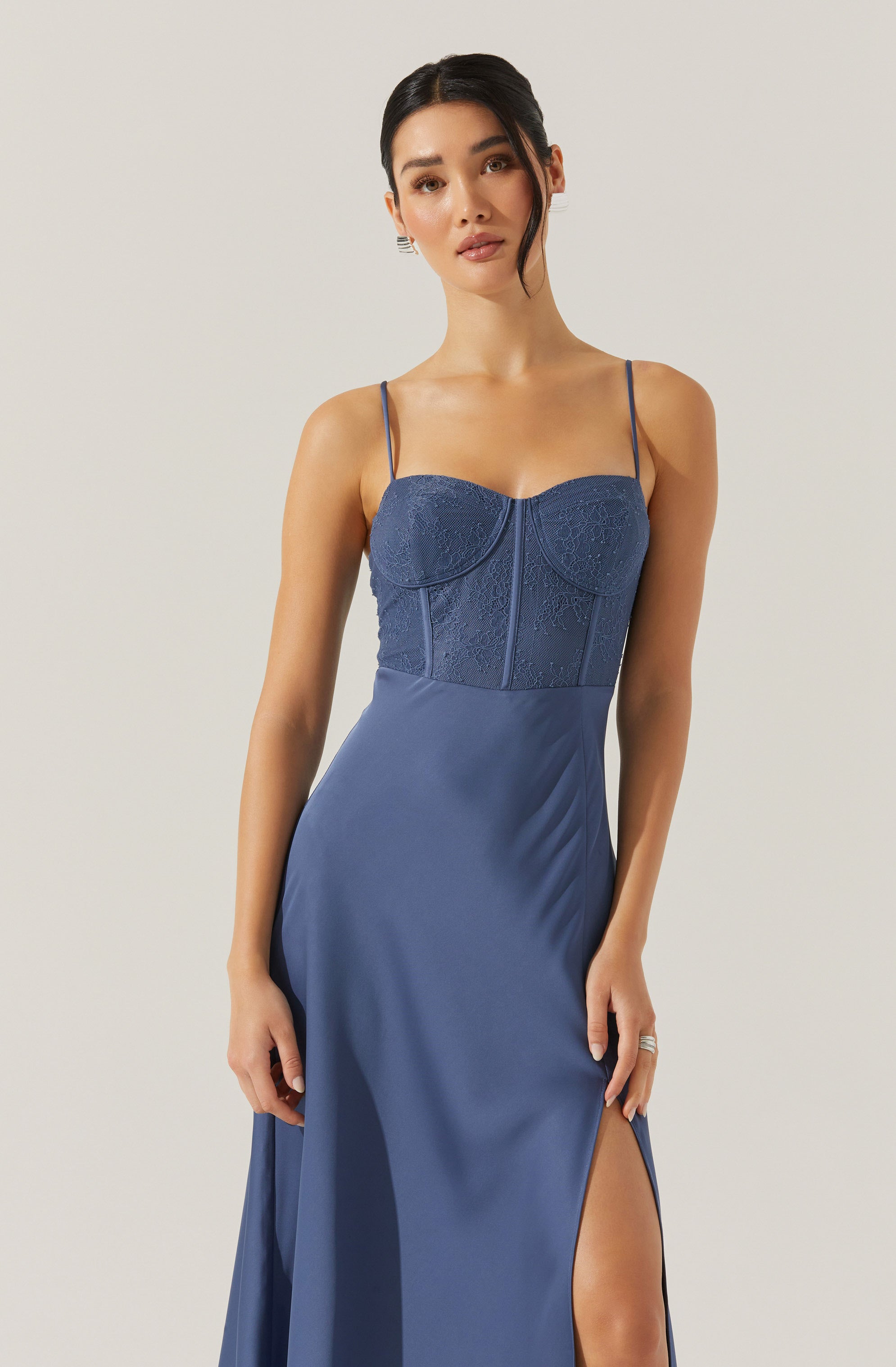 A woman is wearing a blue evening gown featuring a fitted bodice with intricate lace and embroidery details. The dress has thin spaghetti straps and a thigh-high slit on one side.