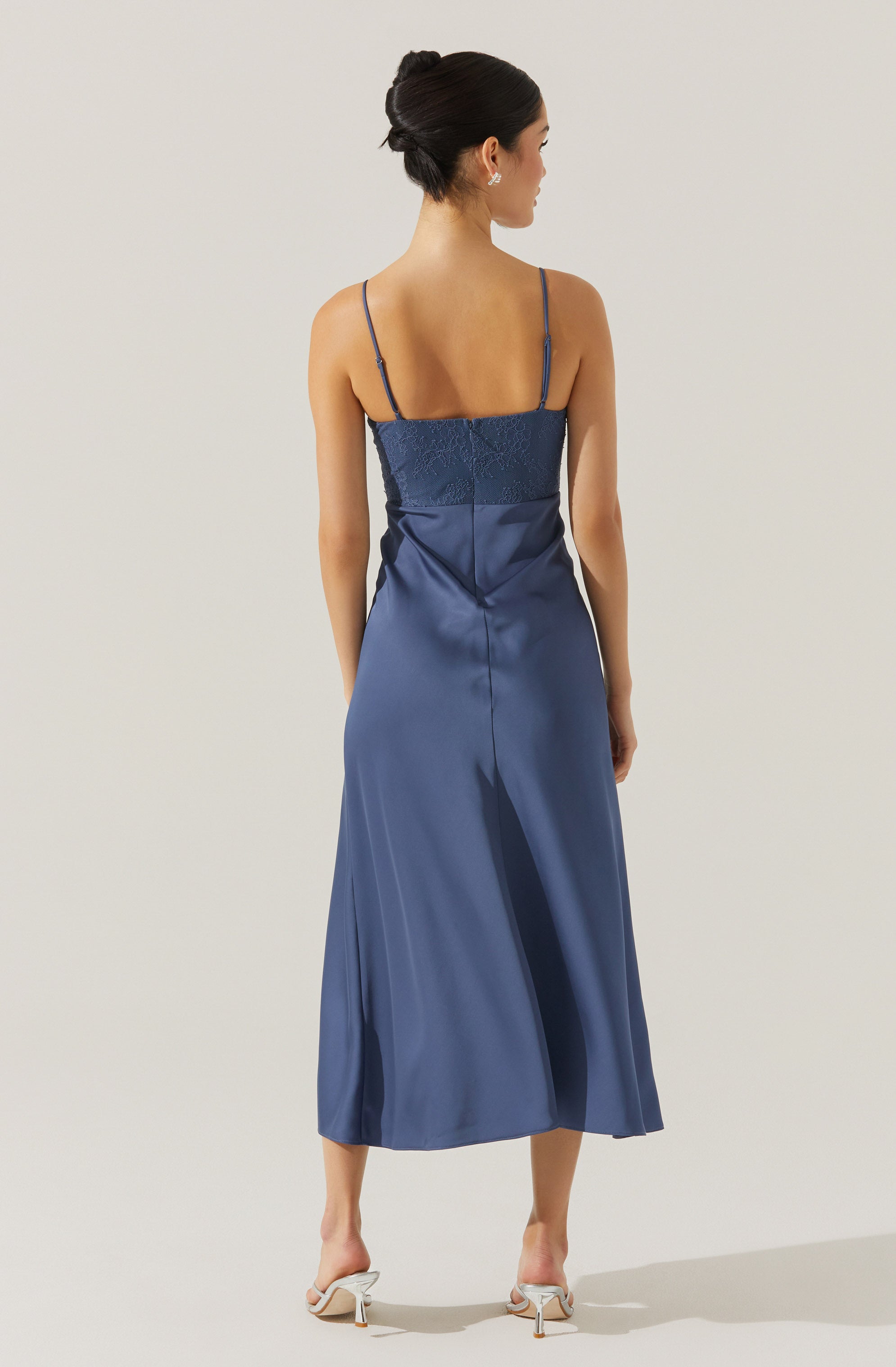 A woman is wearing a navy blue, satin midi dress with spaghetti straps. The top of the dress features a textured, lace-like section, while the skirt is smooth and flowing, reaching mid-calf. She is also wearing light-colored high-heeled shoes. Her hair is styled in an elegant updo.